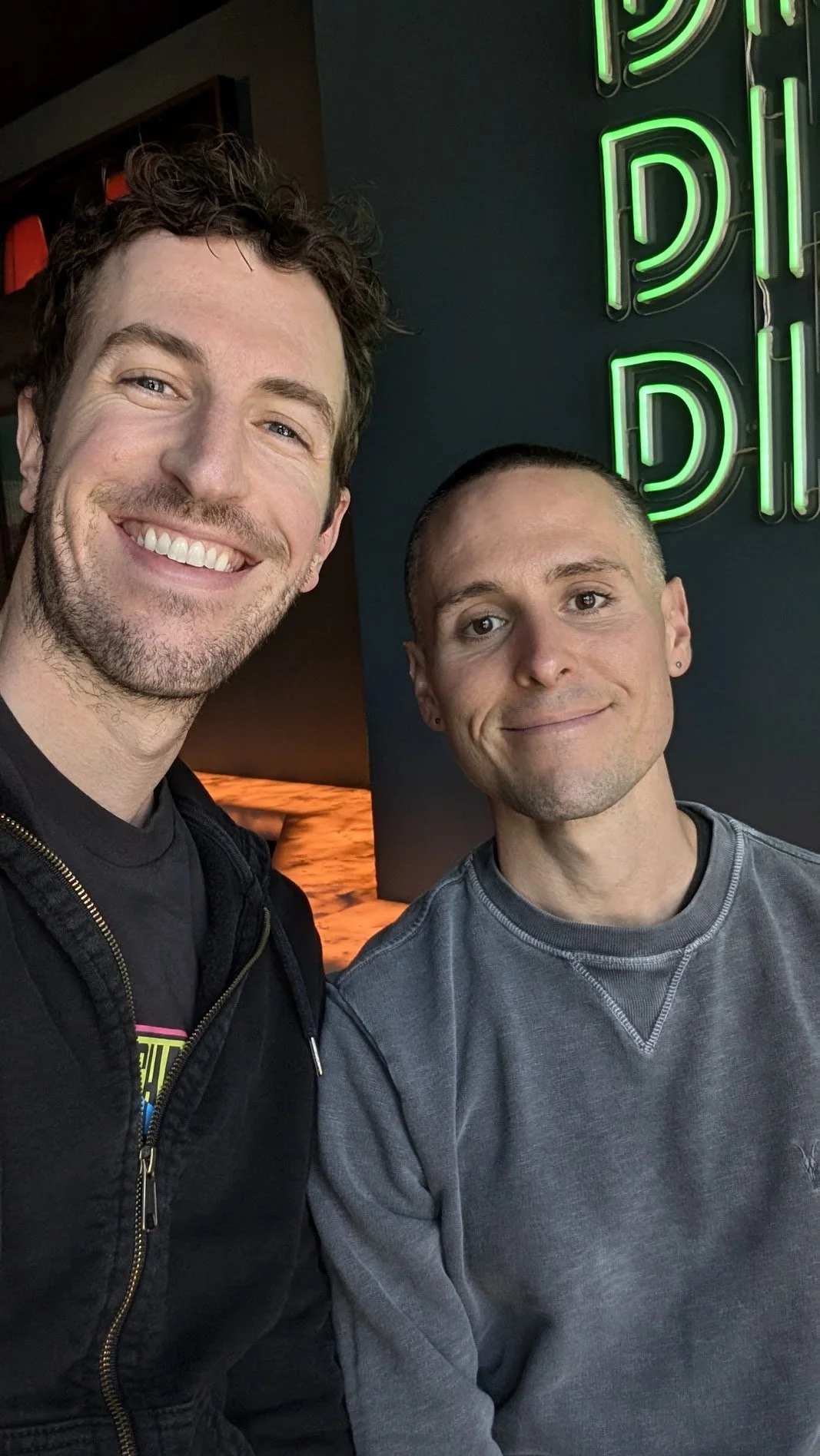 Two men smiling - husbands Carter and Nick Gibson - and taking a selfie in front of a green neon sign in a dimly lit indoor setting.