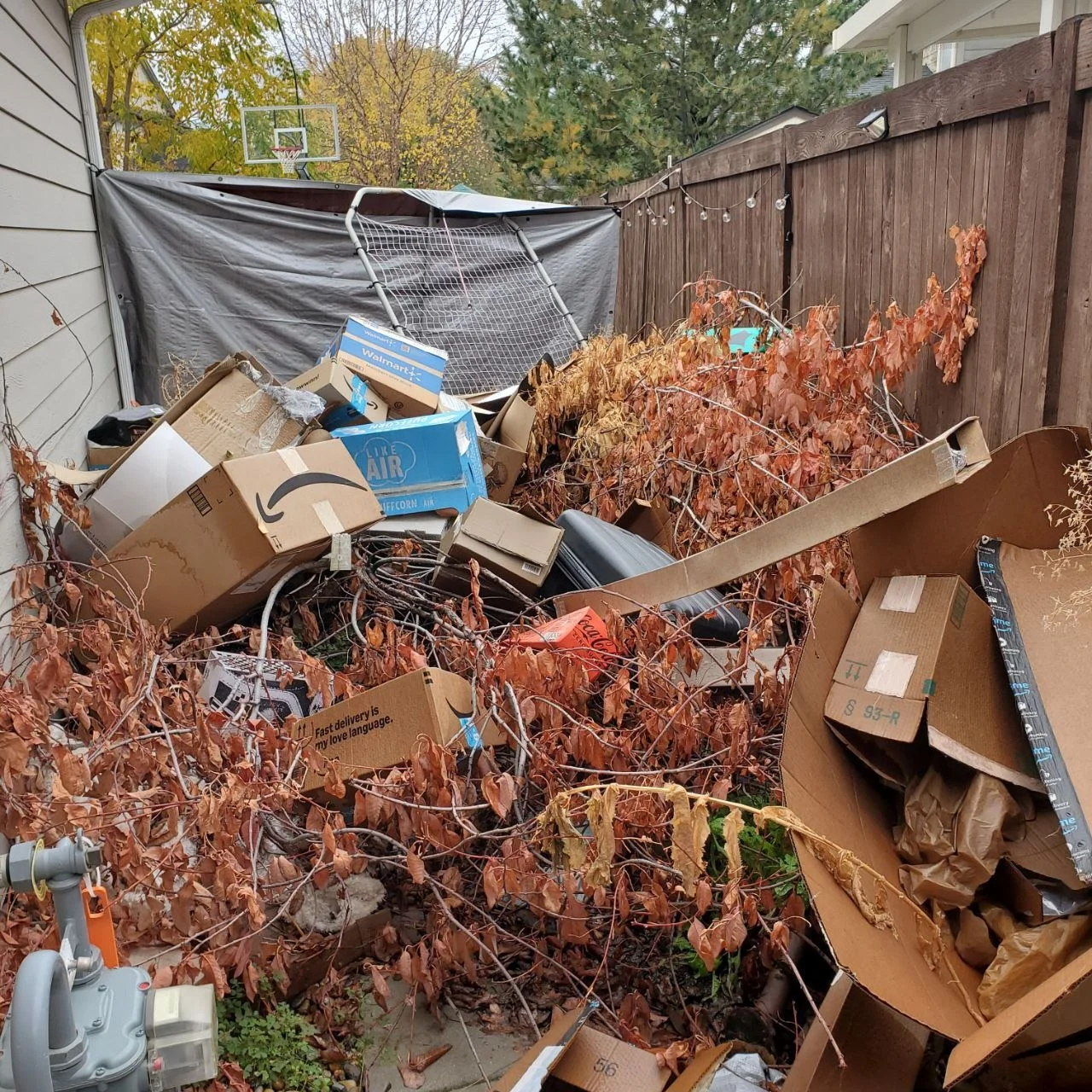 Overflow of discarded cardboard boxes, branches, and miscellaneous trash in a backyard with a wooden fence and string lights.