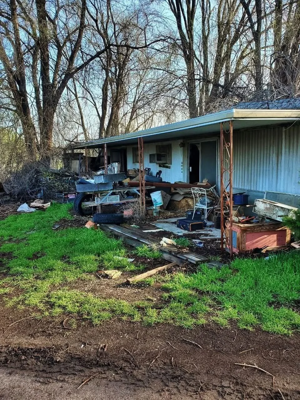 The image shows the front of a house with a damaged porch, cluttered with debris, scattered belongings, and outdoor furniture, surrounded by leafless trees and a mix of grass and dirt ground.