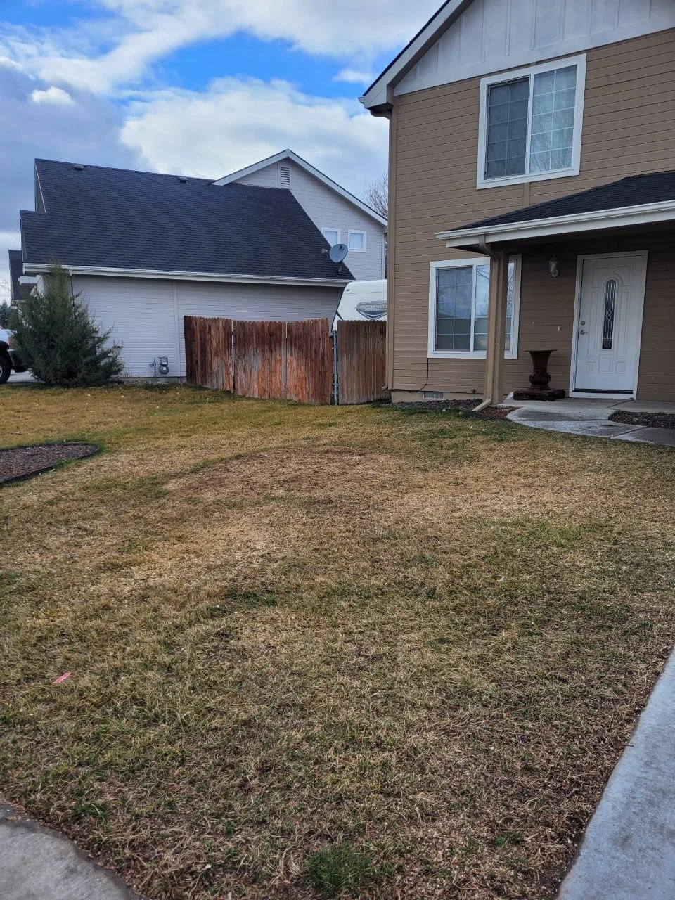 View of a backyard lawn with a brown two-story house on the right and a white house in the background, fenced yard, cloudy sky.
