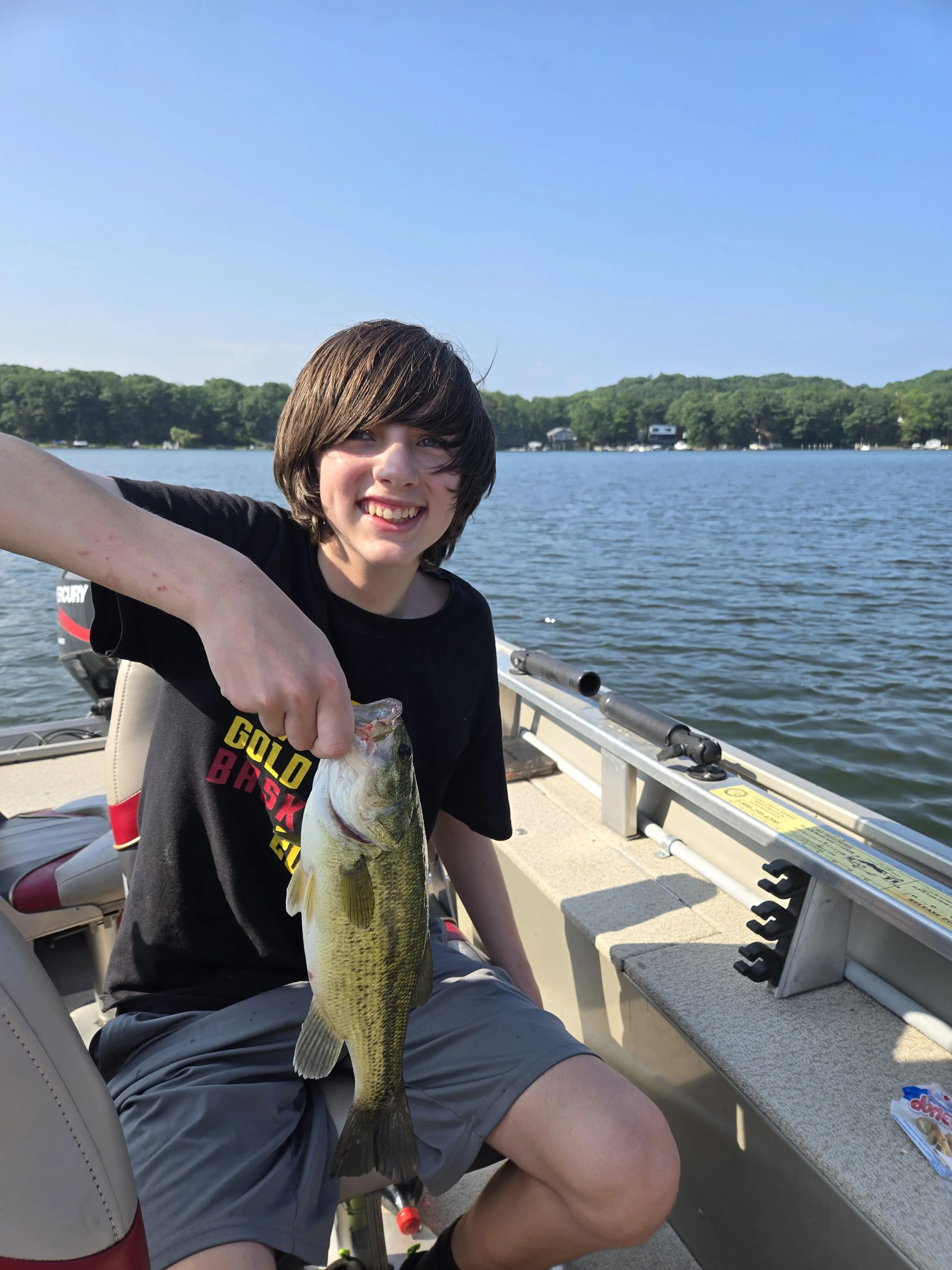 A young person with brown hair smiling and holding a large fish on a boat by a lake, with trees and houses in the background.