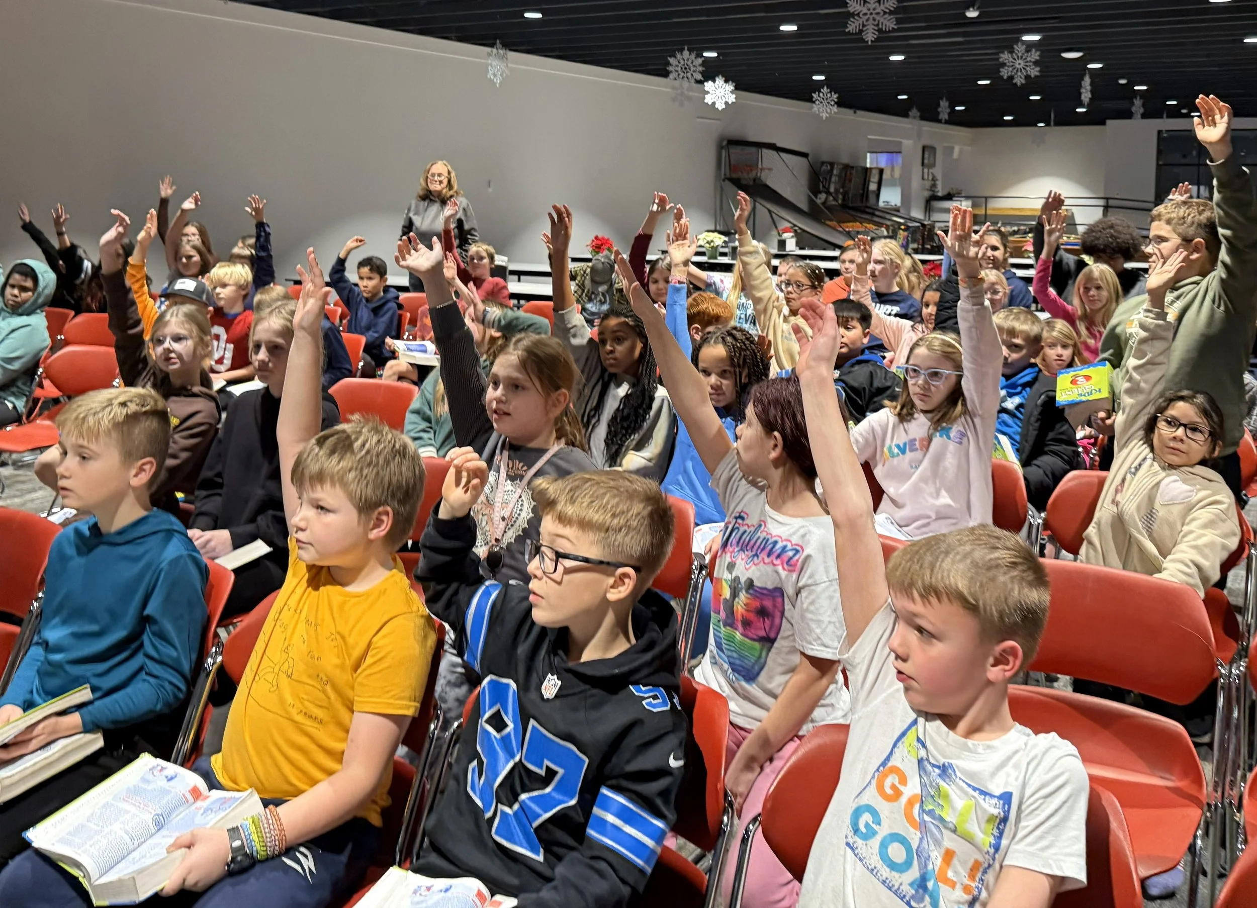 A group of children sitting in chairs, many raising their hands, at upward bound
