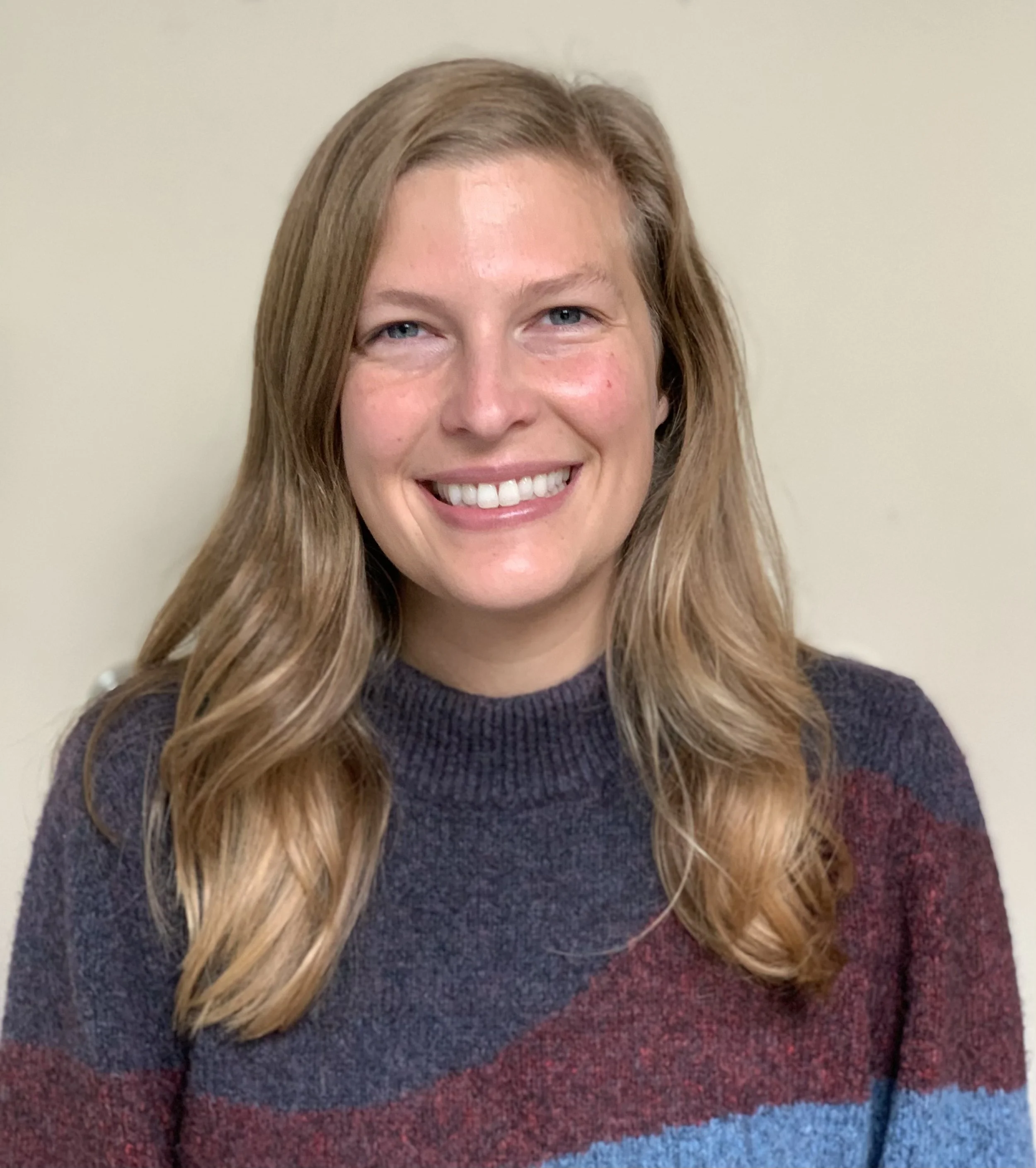 Close-up image of a smiling woman with long, wavy, blonde hair, wearing a dark-colored sweater.