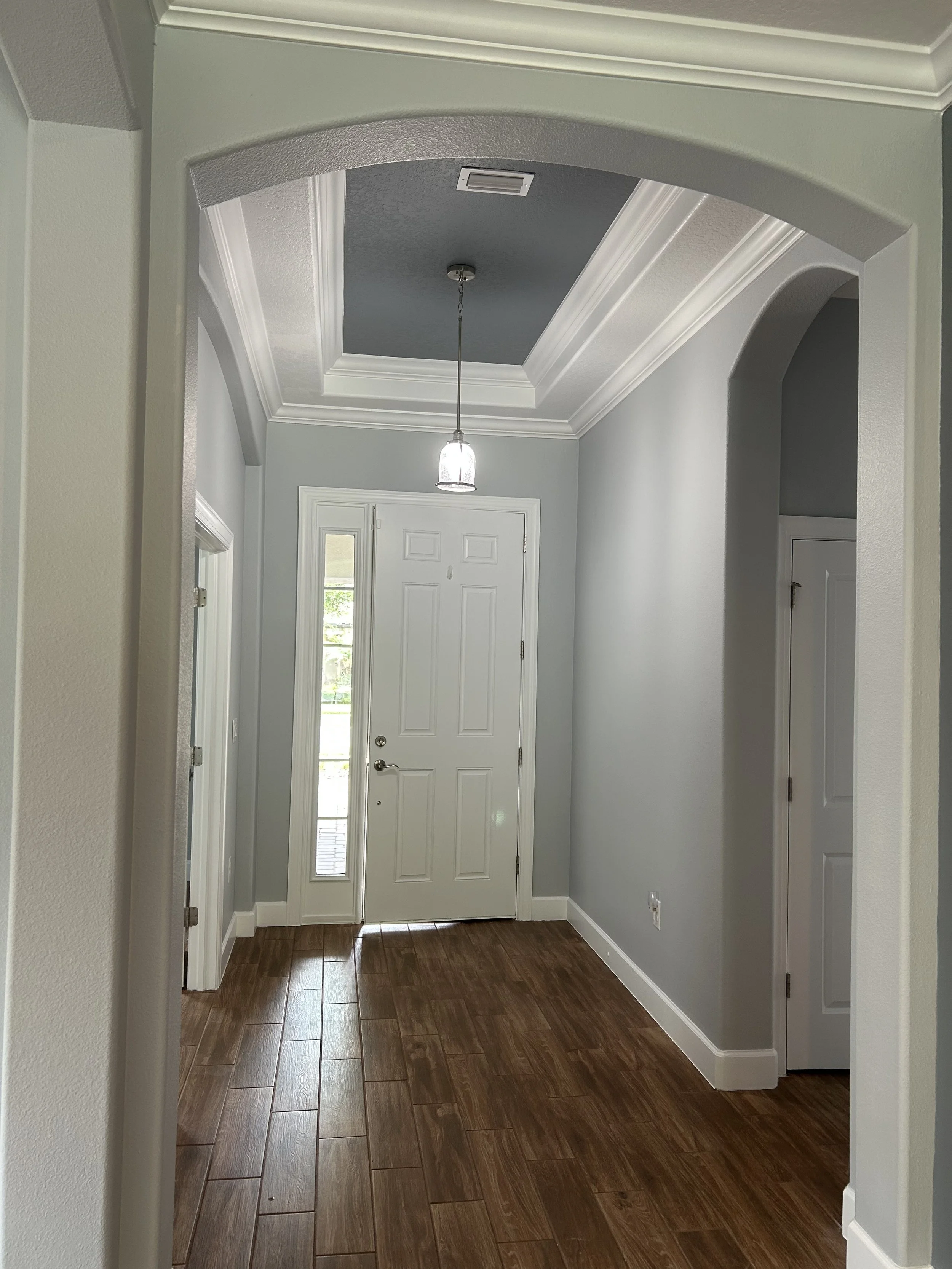 The image shows a home entryway with wood-look tile flooring, white walls, and a white front door with sidelights. An overhead light fixture hangs from the ceiling, which has crown molding and a textured ceiling.