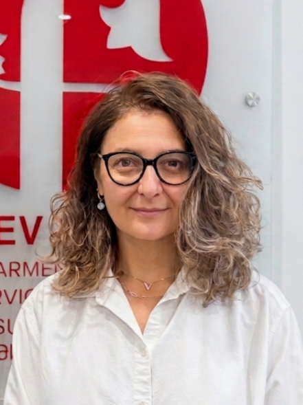 A woman with wavy brown hair, glasses, and a white shirt standing in front of a Red Cross logo.