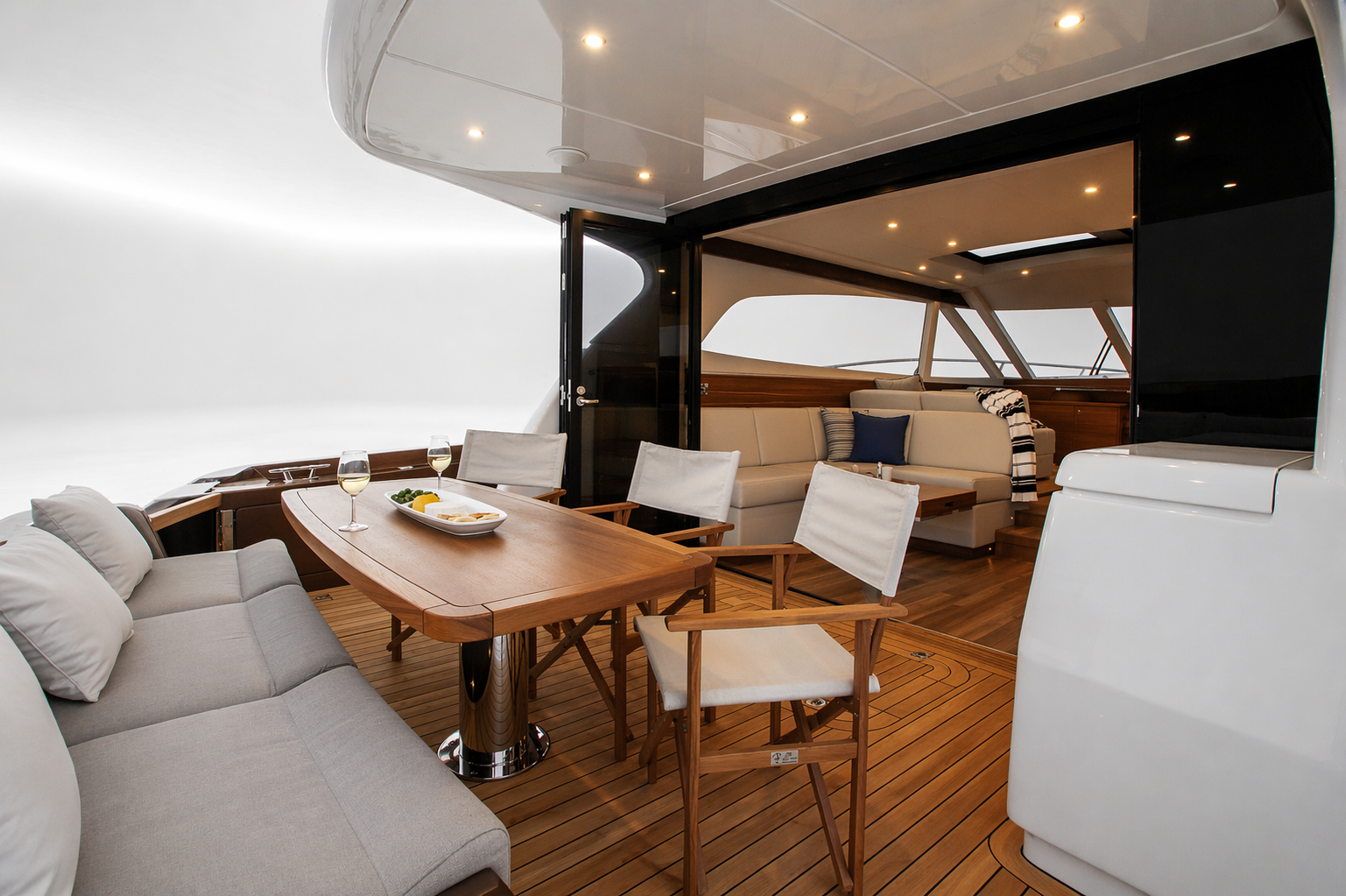 Interior of a yacht with a dining area, featuring a wooden table with two glasses of white wine and a plate of appetizers, surrounded by chairs and a sofa, with large windows and wooden flooring.