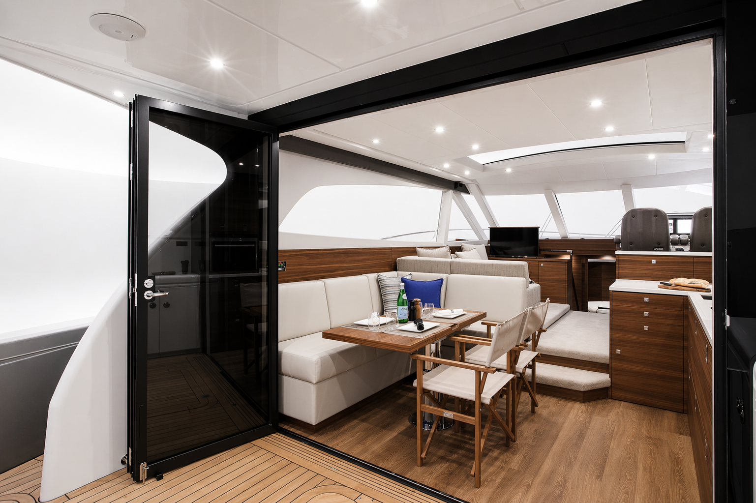 Interior of a modern yacht with a dining area, beige sofa, wooden table, and chairs, with a control station visible in the background.