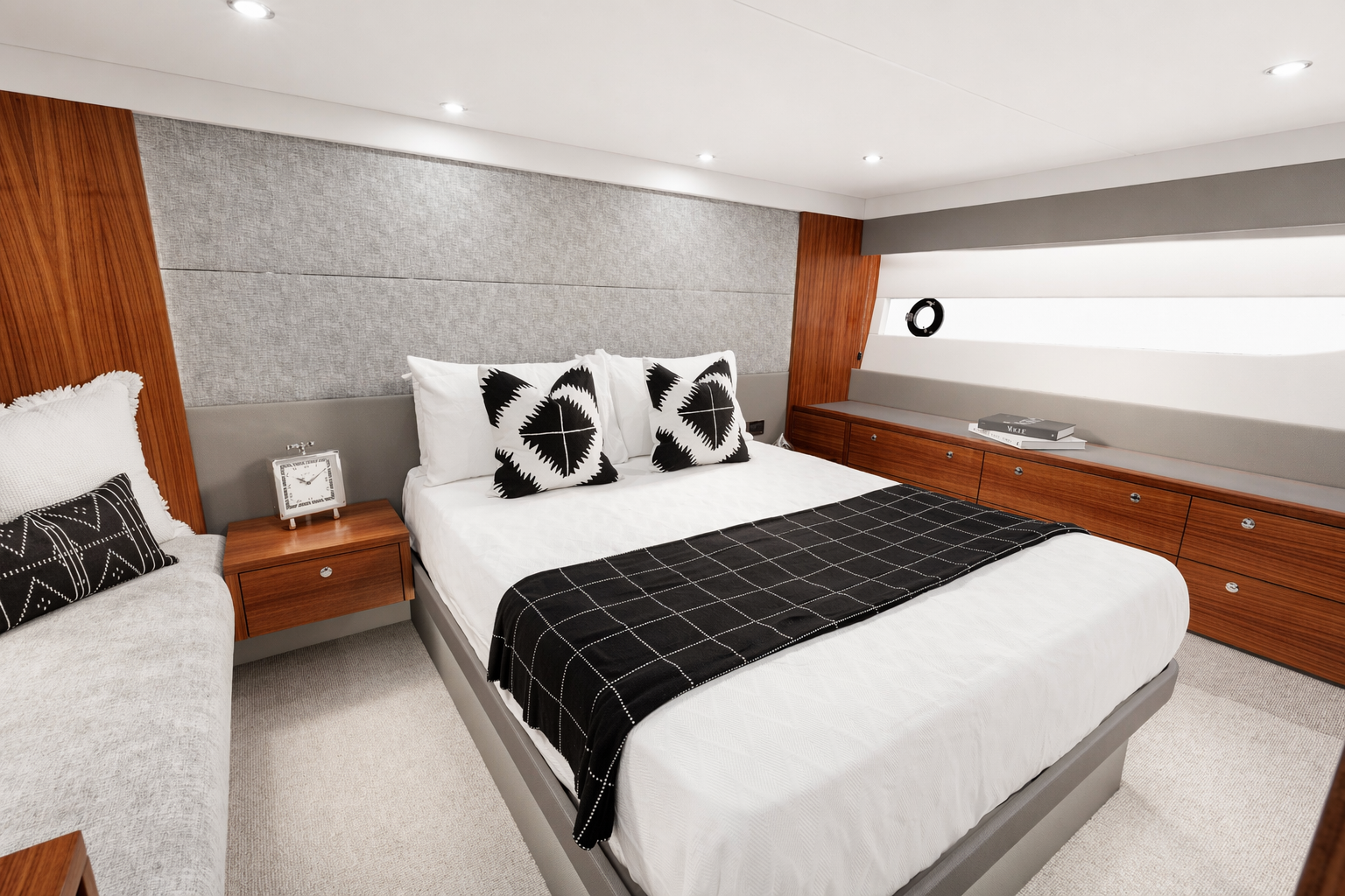 Modern bedroom with king-sized bed, black and white pillows, gray wall, wooden accents, side table with clock, built-in storage, and window with white blinds.