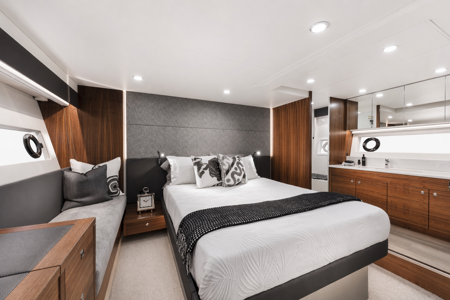 Modern bedroom with a large bed, gray upholstered headboard, decorative pillows, wooden accents, a built-in side sofa, and a vanity with mirror and accessories.