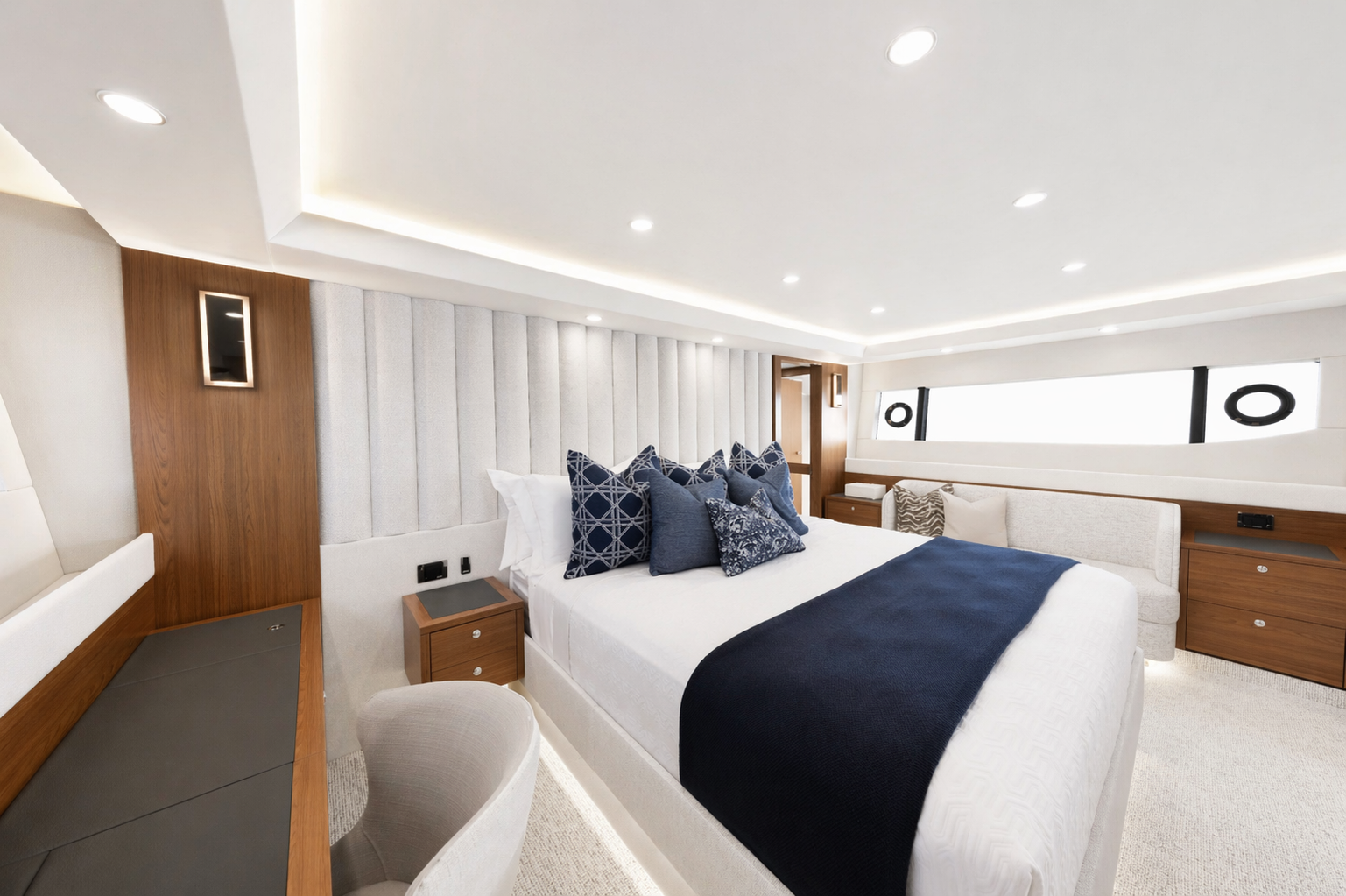 Luxury hotel bedroom with white bed, navy blue throw blanket, four navy blue pillows, beige armchair, wooden side tables, and modern decor with circular windows.