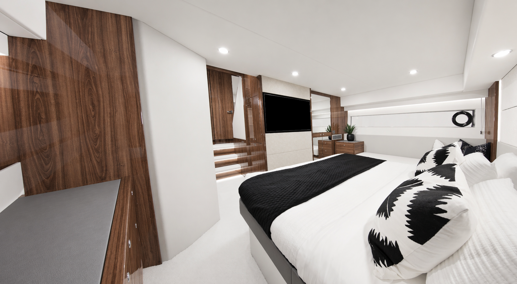 Modern bedroom with white walls, a wooden accent wall, a large bed with white bedding and black decorative pillows, a black throw blanket, and a wall-mounted flat-screen TV. There are two potted plants on a wooden nightstand and recessed ceiling lights.
