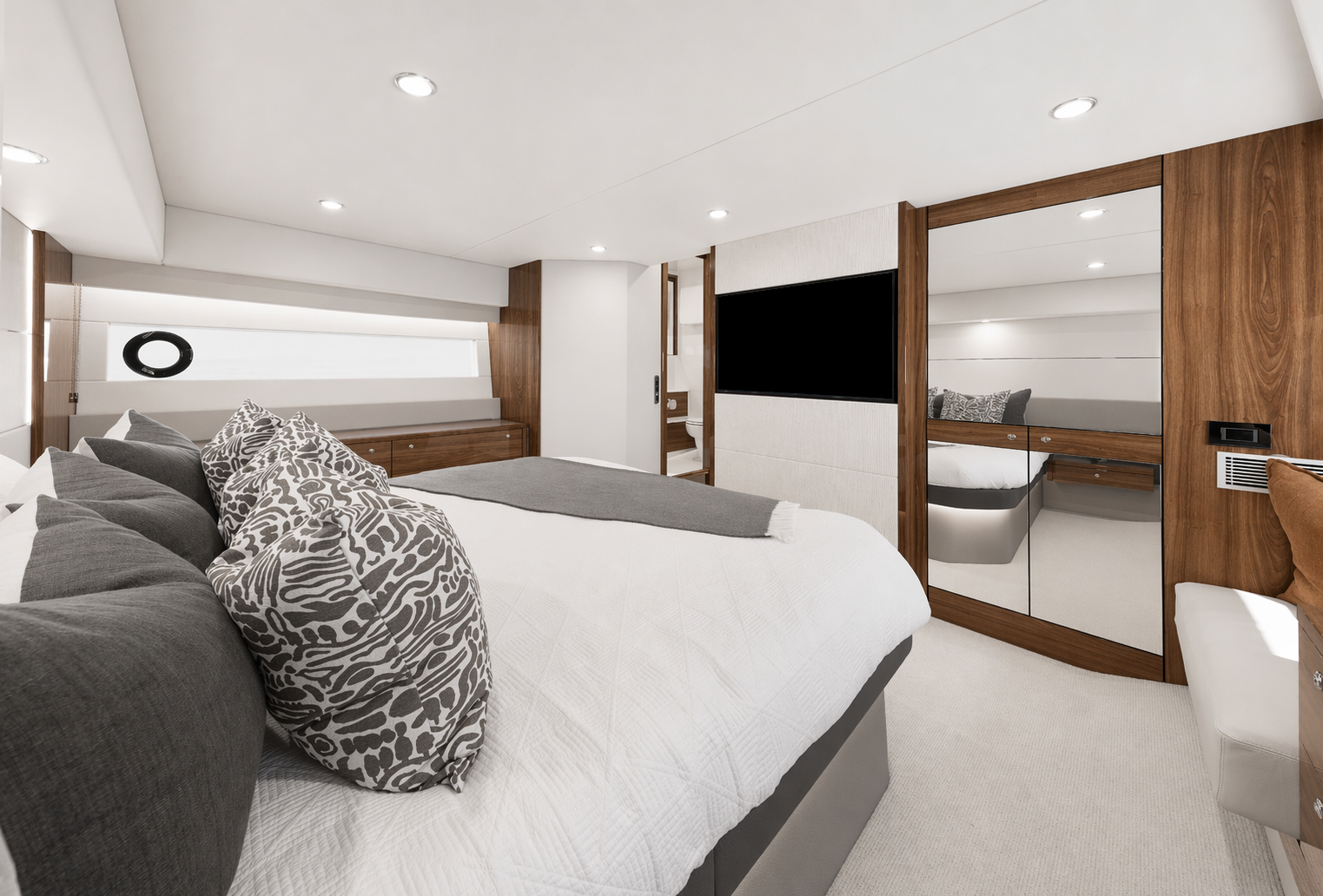 Modern yacht bedroom with a white bed, gray and patterned pillows, wooden accents, mirrored closet, and a mounted TV.