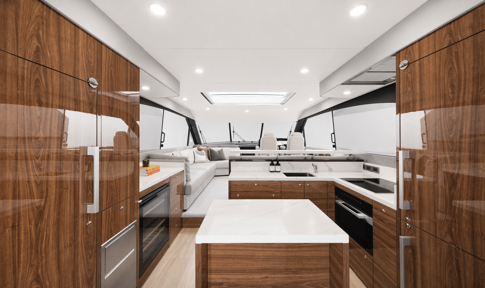 Modern yacht interior kitchen with wood cabinetry, white countertops, and built-in appliances, with a living area and large windows at the front.