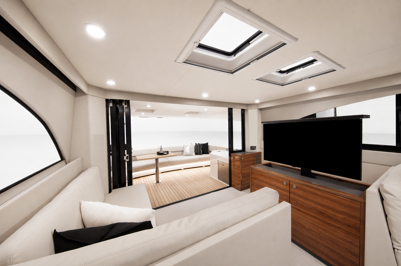 Interior of a modern yacht living room with white sofa, flat-screen TV on wooden cabinet, and sliding glass door leading to balcony with seating area. Ceiling has two skylights.