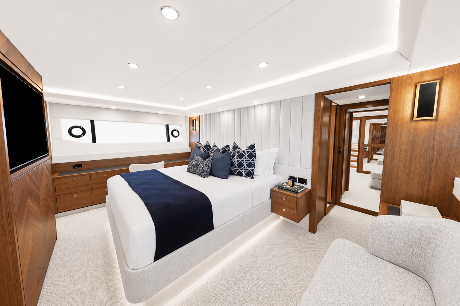 Modern bedroom with white bed, navy throw blanket, multiple navy and patterned throw pillows, side table with books and phone, wood panel walls, and a private sitting area visible through a glass door.