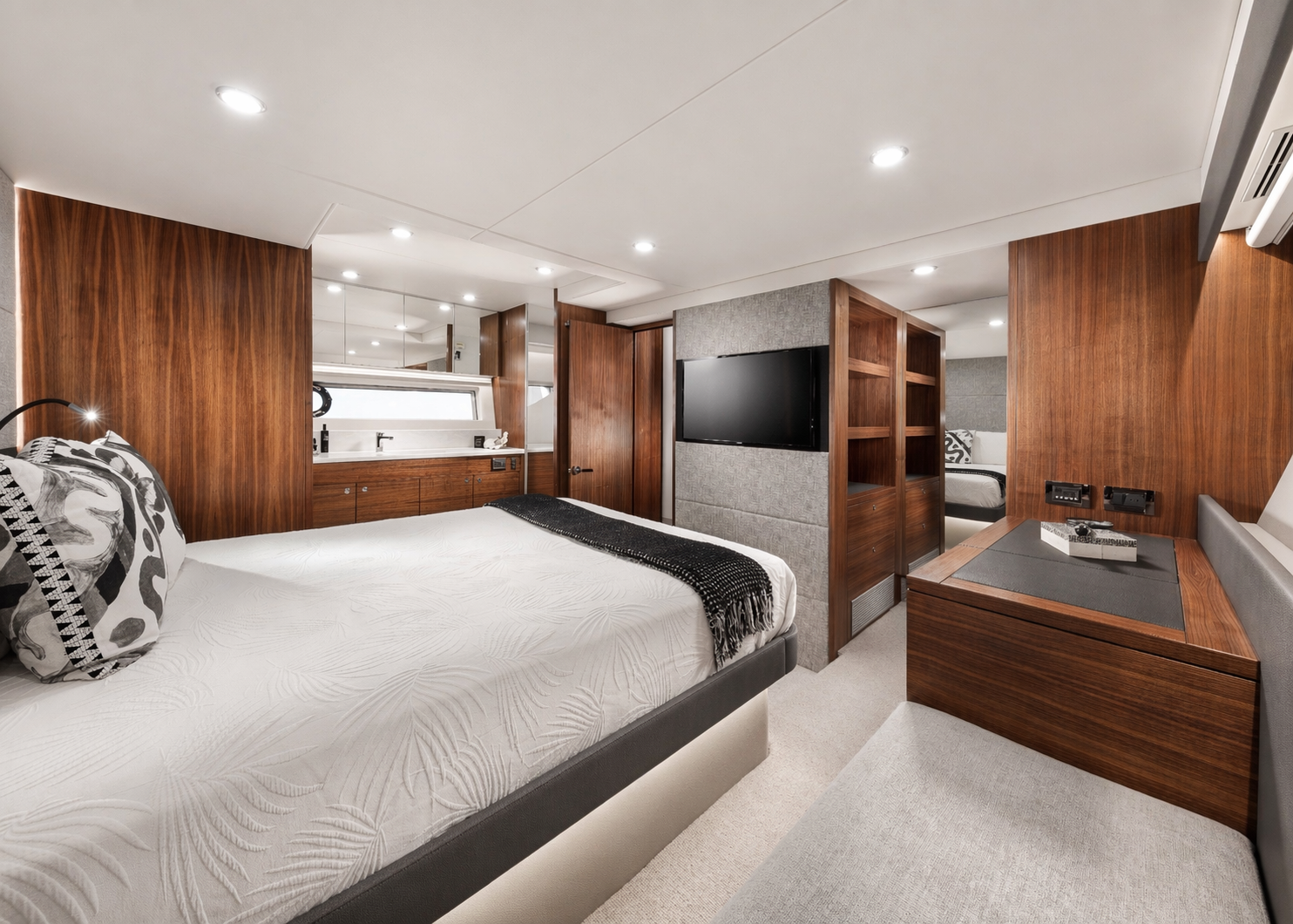 Interior of a modern bedroom with a white bed, black and white pillows, wooden panels, a flat-screen TV, and a small workspace with a desk and seating.