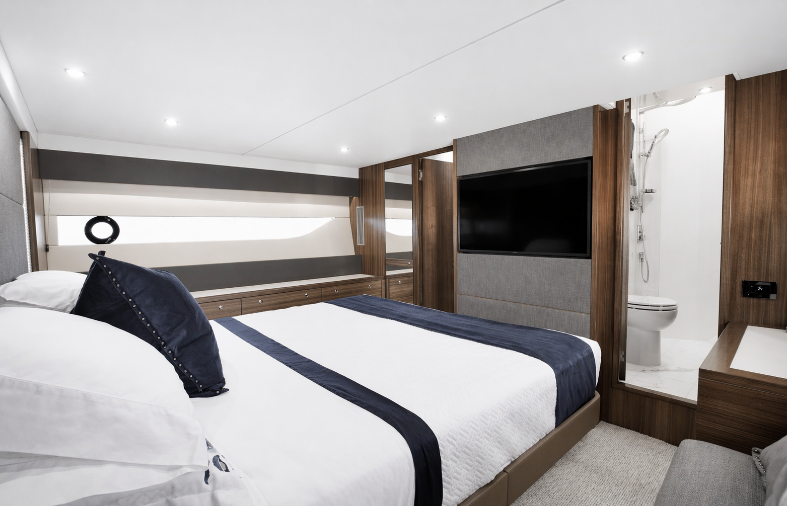 Maritimo motoryacht with a bed featuring white and navy bedding, navy pillows, built-in wooden cabinetry, a wall-mounted TV, and an open bathroom with a shower and toilet visible in the background.