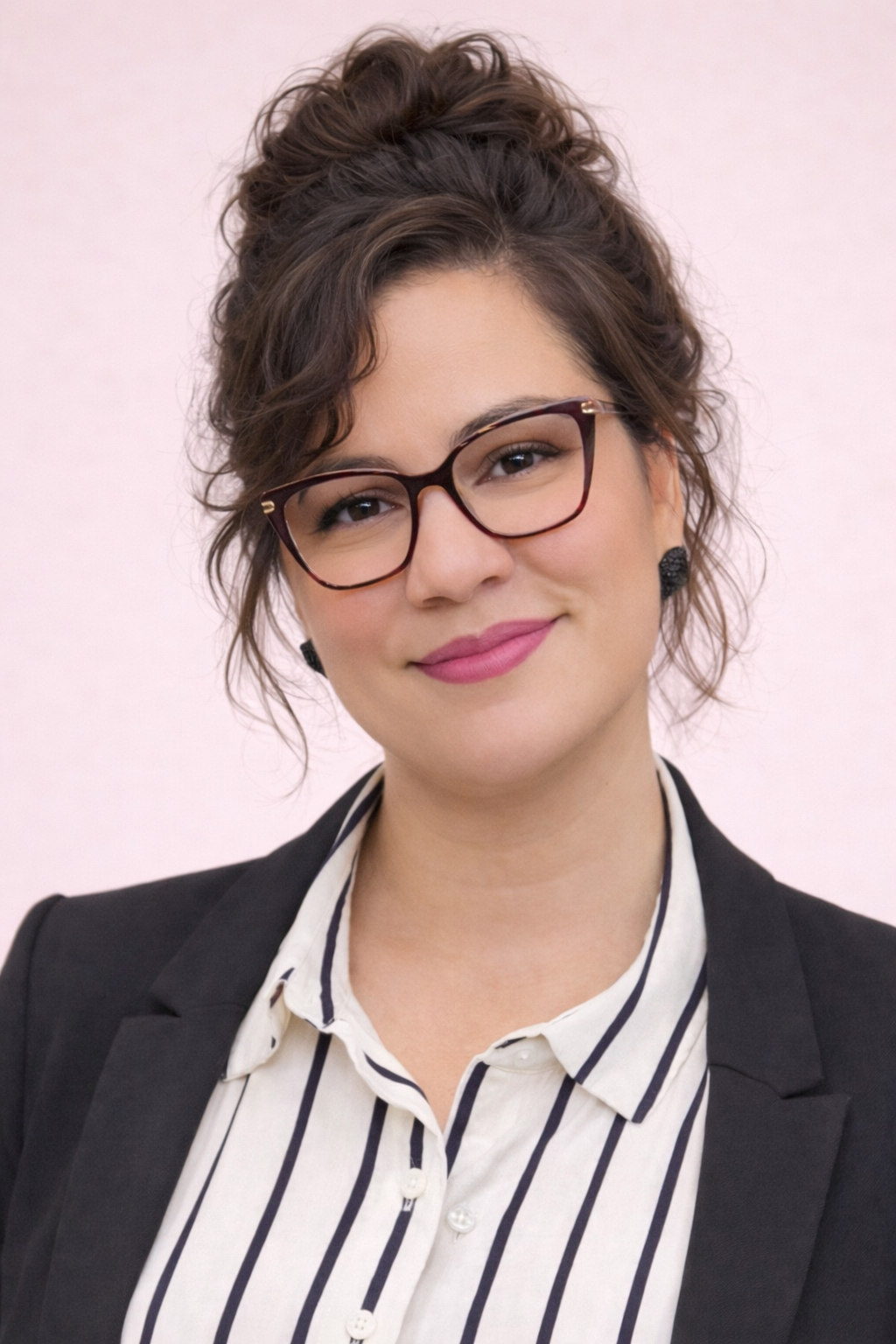 A woman with dark, curly hair tied up in a bun, wearing glasses, black earrings, a black blazer, and a white blouse with black vertical stripes, smiling against a plain light pink background.