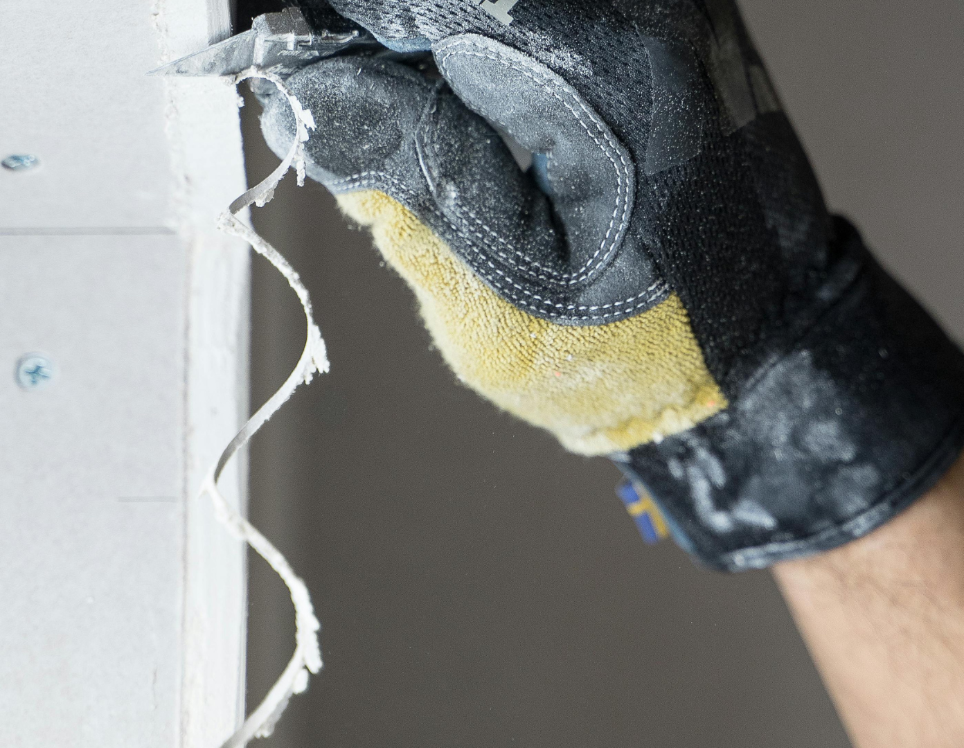 Close-up of a black work glove with yellow padding being used to drill into a gray wall, with drywall dust and torn drywall material around the drill hole.