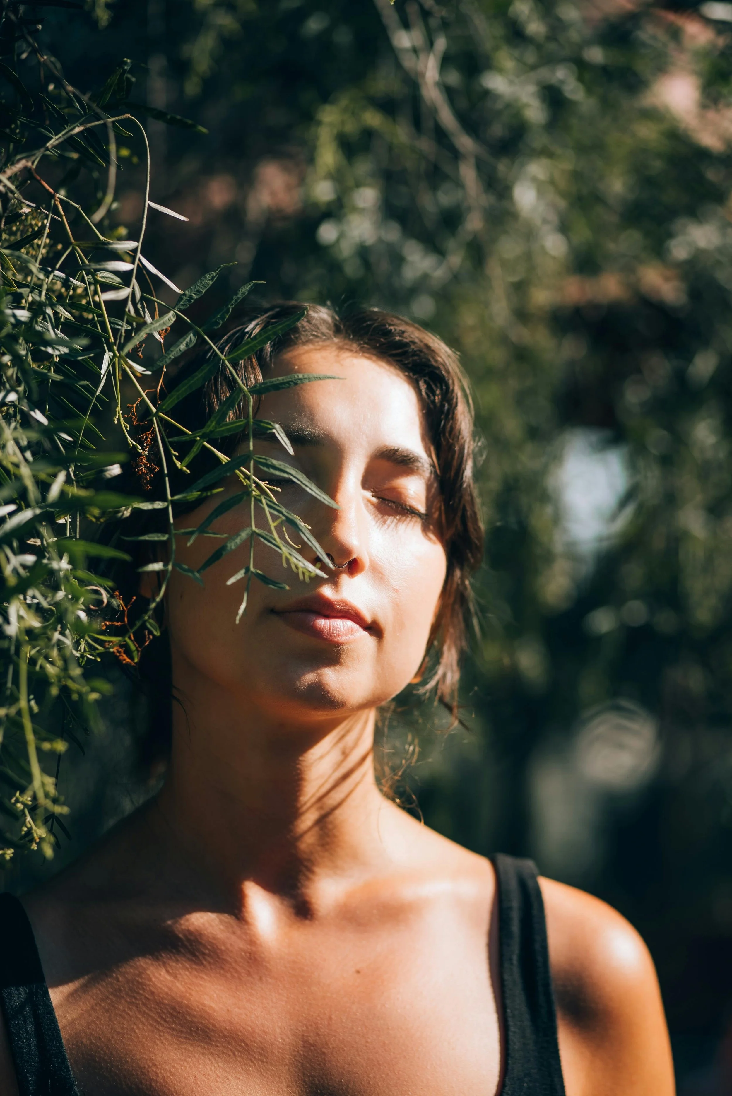 A woman stands with eyes closed and a serene expression, her face bathed in warm sunlight amid the trailing branches of a leafy tree.