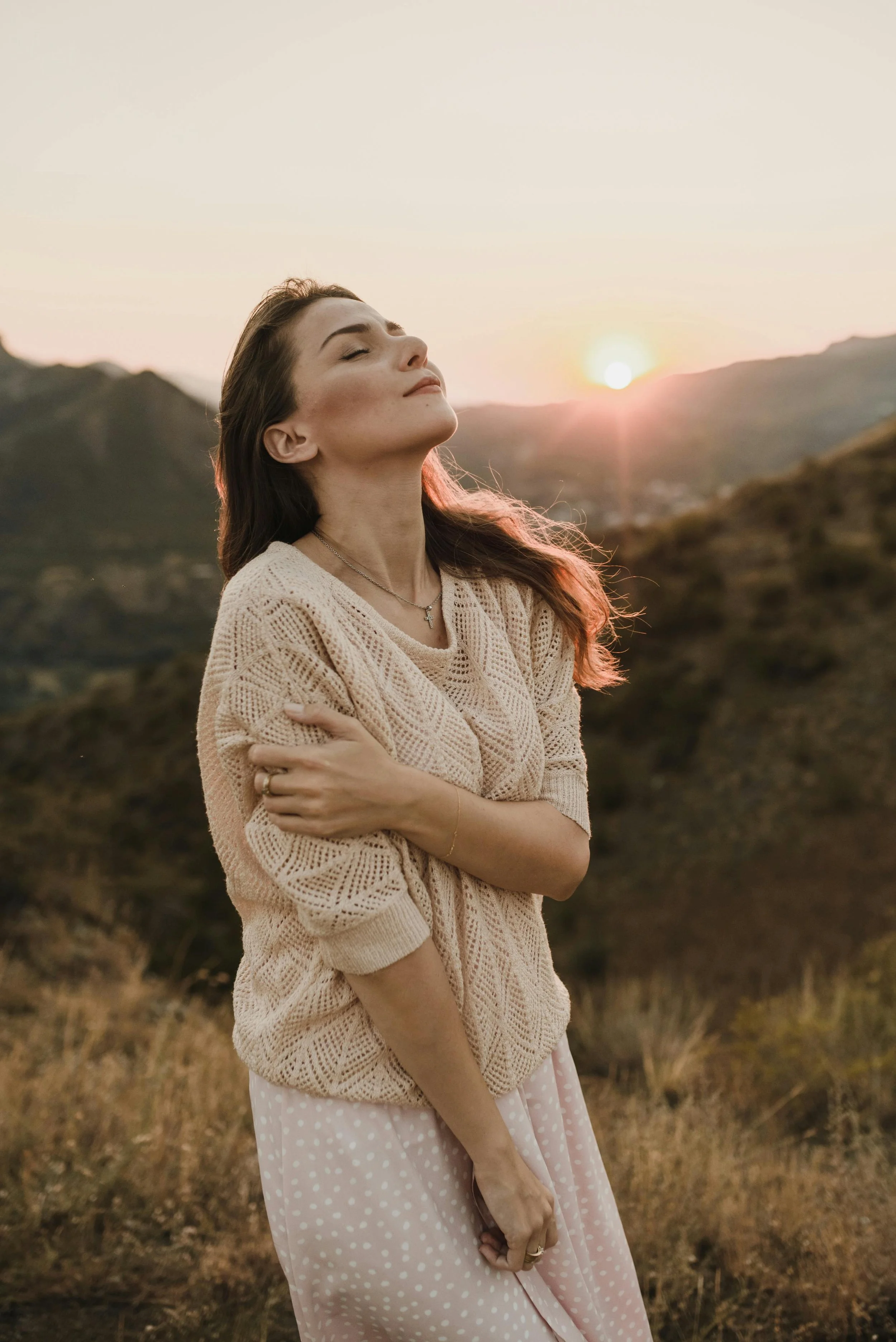 A woman tilts her face upward with eyes closed, arms wrapped around herself, wearing a cream knit sweater and floral skirt, standing in a golden sunset landscape with mountains behind her.