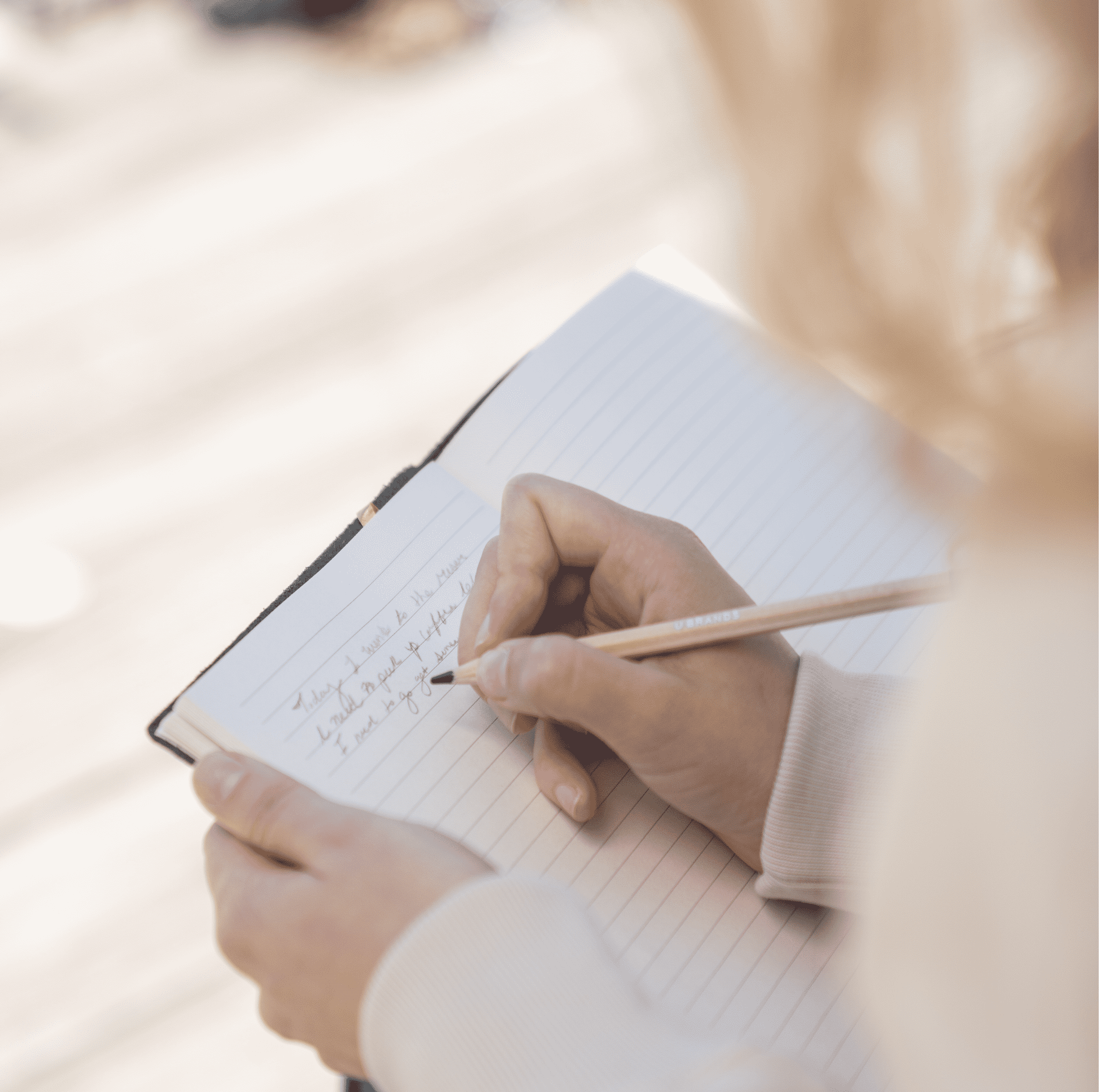 A close-up of a hand writing in a lined notebook, a pencil held lightly, in soft warm light.