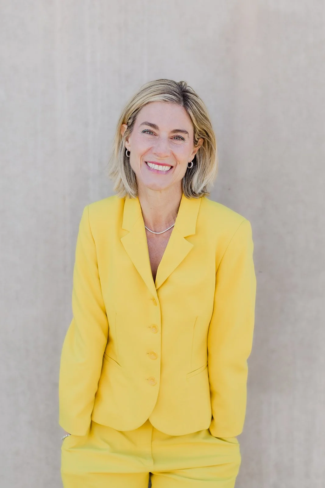 Dr. Kim stands smiling confidently against a neutral concrete wall. She wears a tailored yellow blazer and matching trousers, with a delicate silver necklace and hoop earrings.