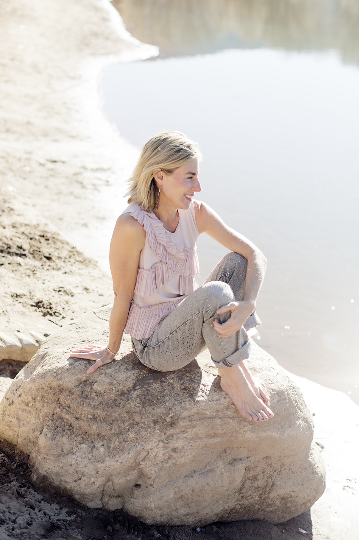 Dr. Kim sits barefoot on a large rock beside a calm shoreline, smiling as she gazes off to the side. She wears a blush ruffled sleeveless top and light gray jeans.