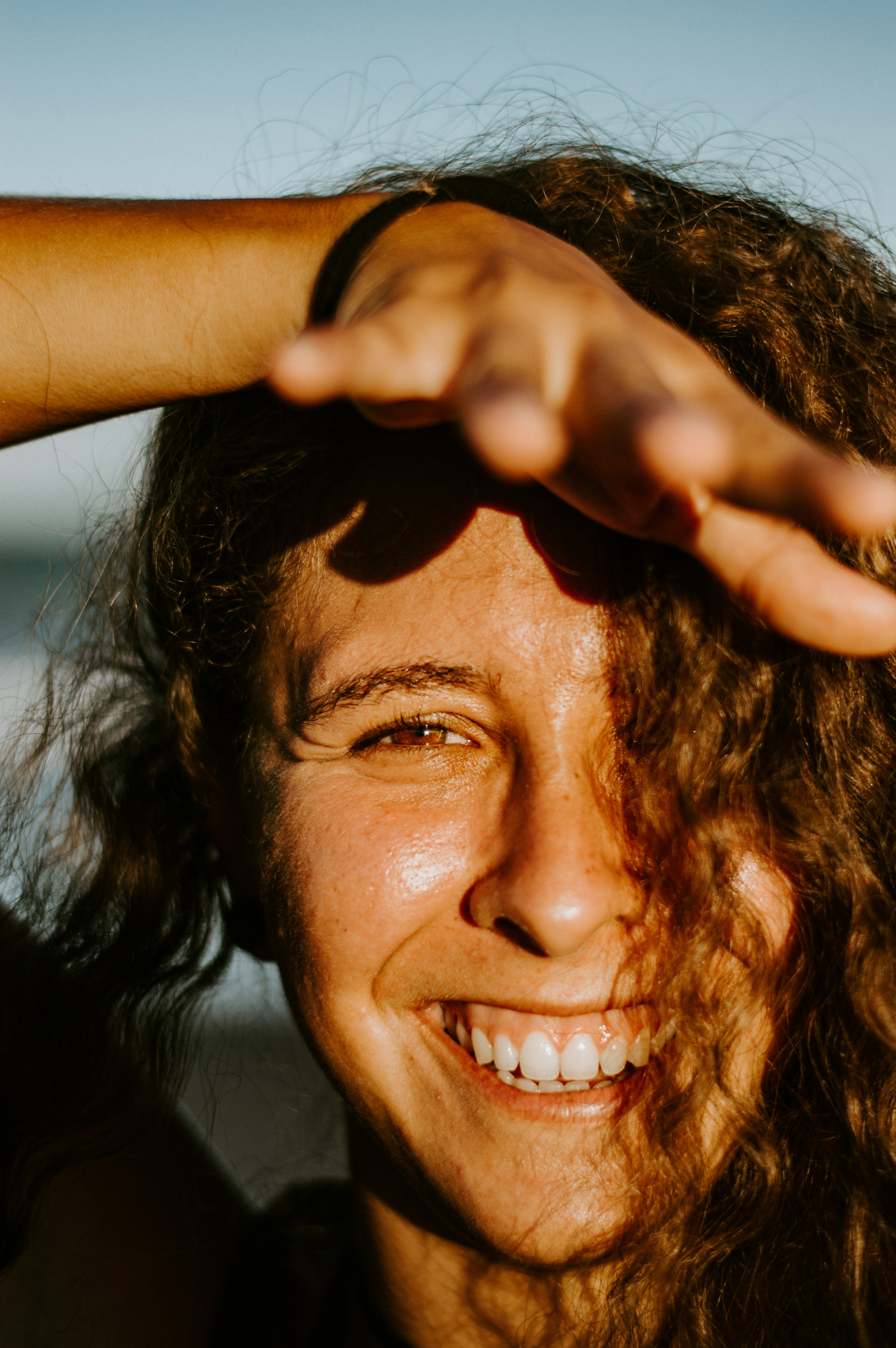 A close-up of a woman with curly hair laughing joyfully, her hand raised above her forehead shielding her eyes from bright sunlight.