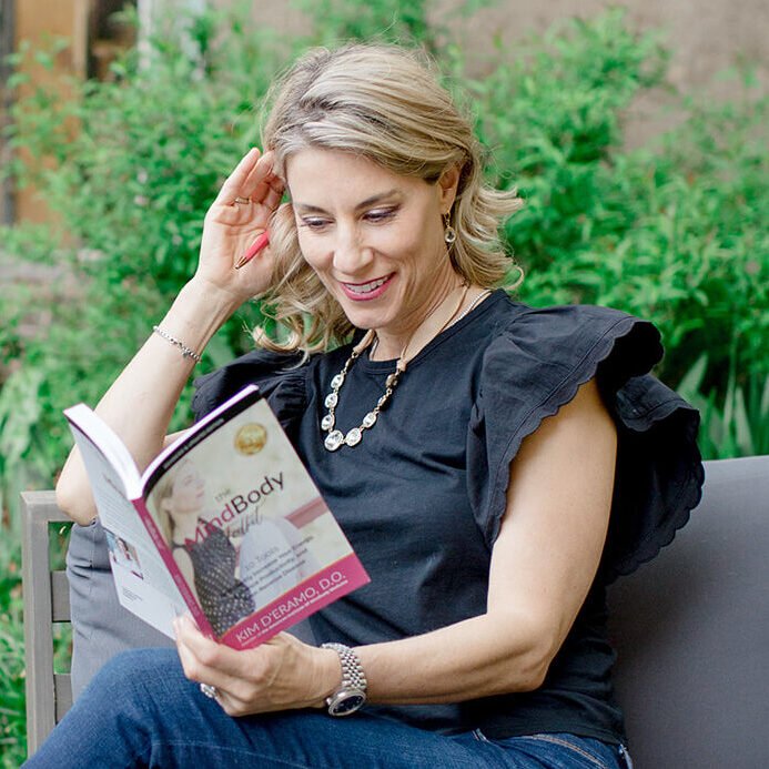 Dr. Kim smiles as she reads a copy of The MindBody Toolkit, seated outdoors amid lush green foliage.