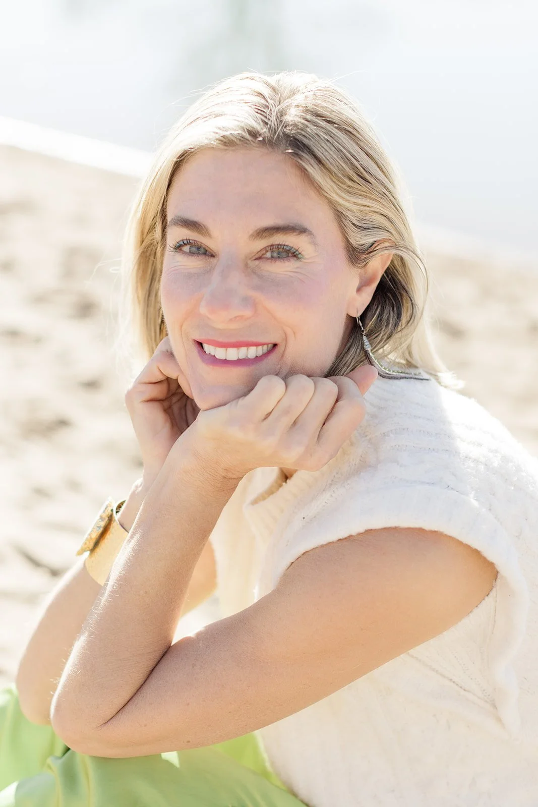A close-up portrait of Dr. Kim D'Eramo smiling warmly at the camera, both hands cupped under her chin, wearing a white knit top and drop earrings, with a softly blurred sandy beach behind her.
