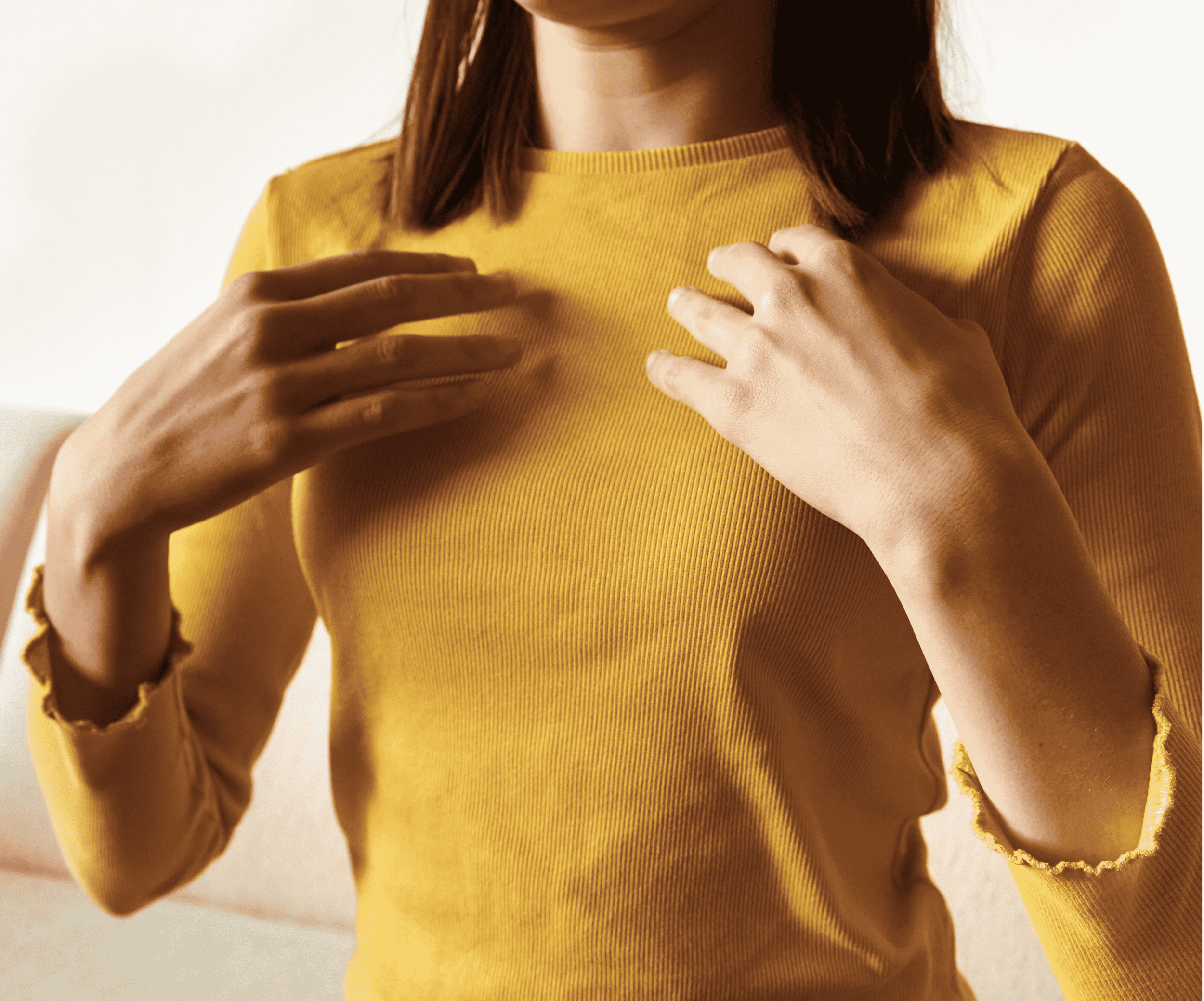 A person places both hands gently on their chest, wearing a mustard yellow ribbed top.