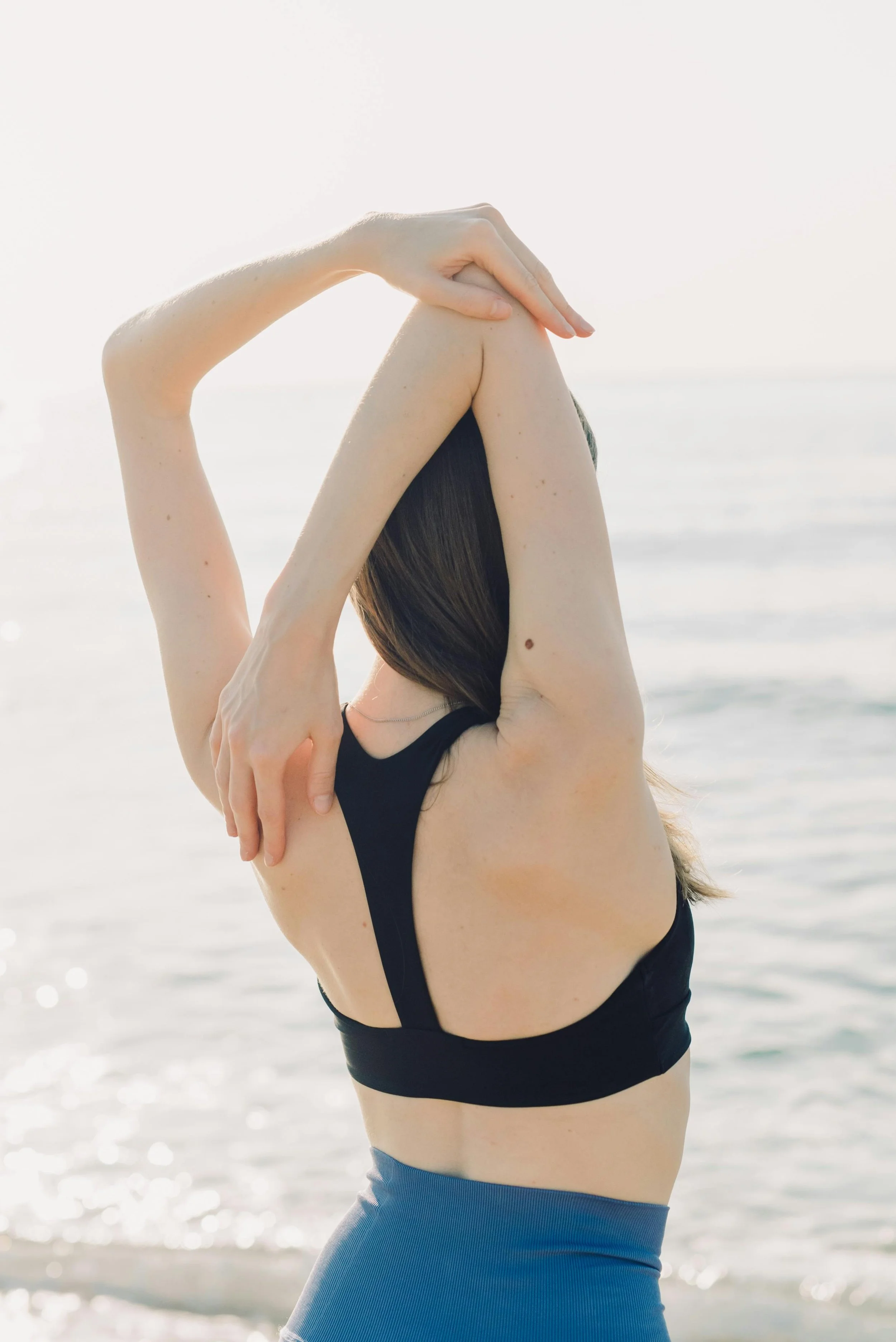 A close-up of a woman in a black tank top stretching her arm overhead near calm, softly lit water.