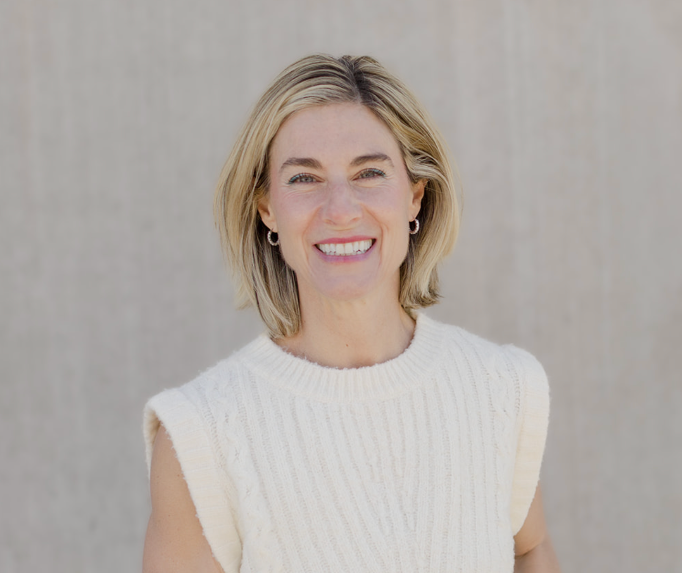 Dr. Kim smiles warmly at the camera, wearing a cream ribbed sweater and hoop earrings, against a soft neutral background.