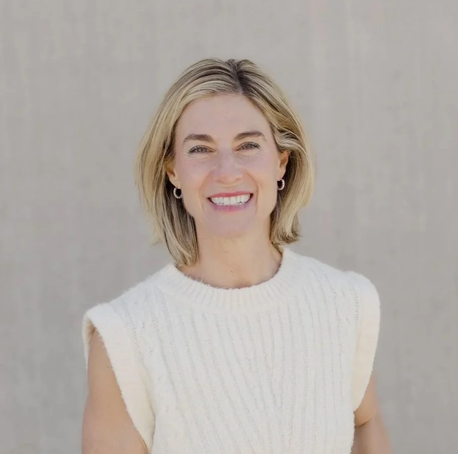 Dr. Kim D'Eramo smiles warmly at the camera, her back against a concrete wall. She is wearing a white sleeveless sweater.