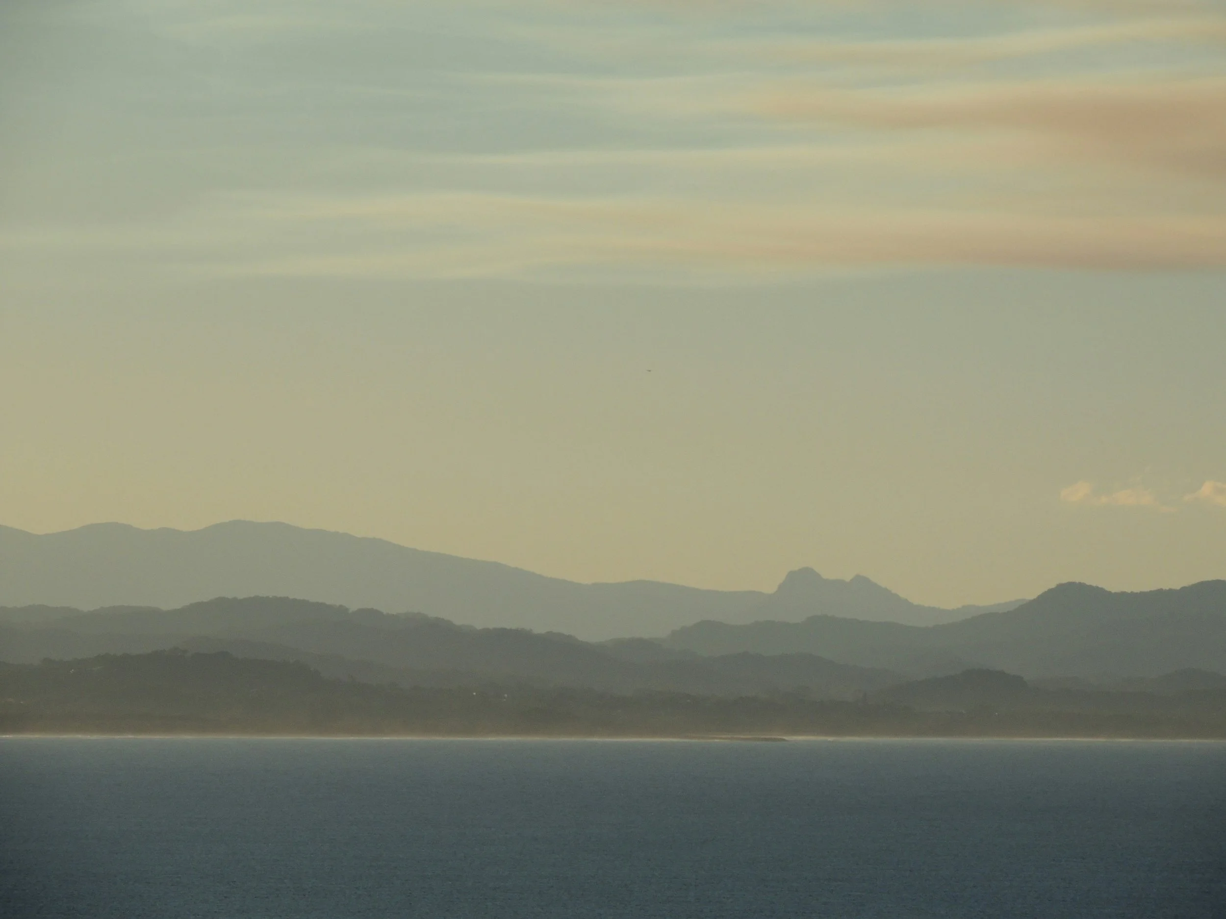 A tranquil coastal landscape at dusk, with calm blue-gray water in the foreground, a distant shoreline, and layered mountain silhouettes fading into a hazy, muted sky with soft peach and blue tones.
