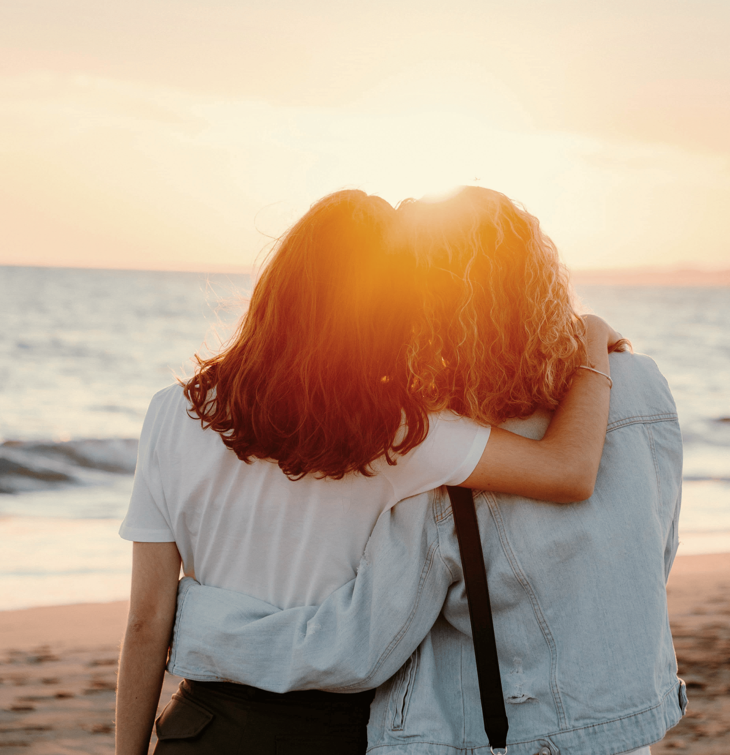 Two people stand with their backs to the camera, arms around each other, facing a calm ocean at sunset.
