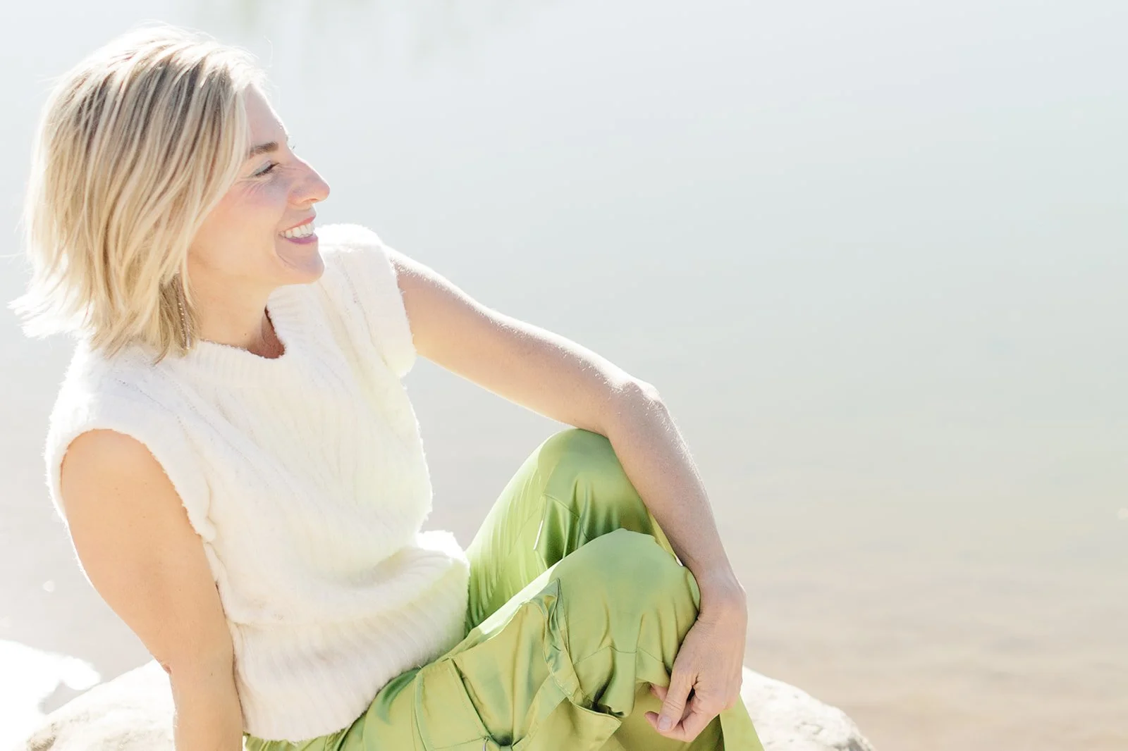 Dr. Kim D'Eramo sits outdoors near water, smiling and looking to the side. She wears a white sleeveless knit top and sage green satin wide-leg pants, with one knee drawn up and her hand resting near her face. The background is softly blurred.