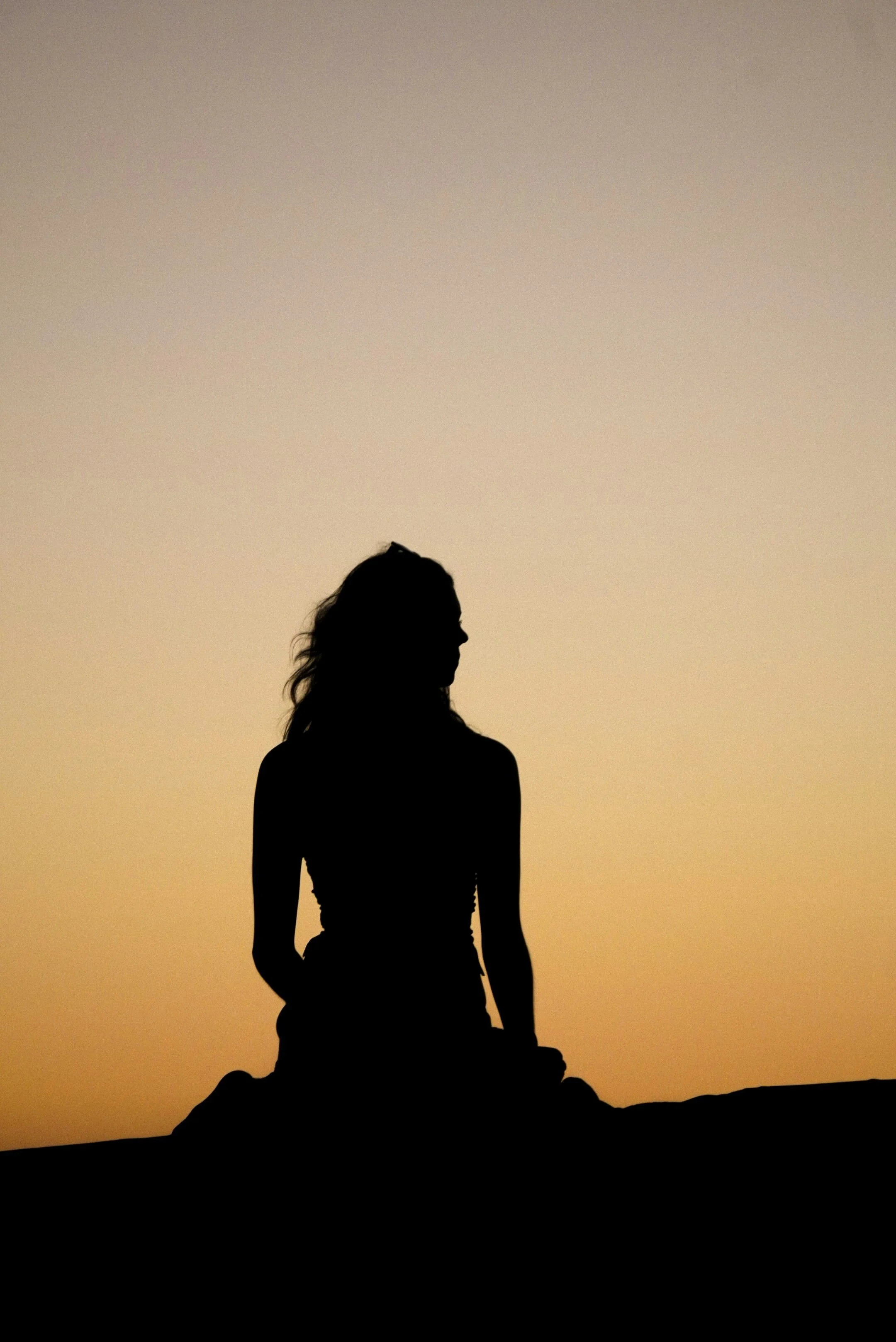 A dark silhouette of a woman with long wavy hair sitting cross-legged on a rock, facing slightly to the right. The background is a warm gradient sky transitioning from soft peach at the horizon to pale gray above, suggesting dusk or dawn.