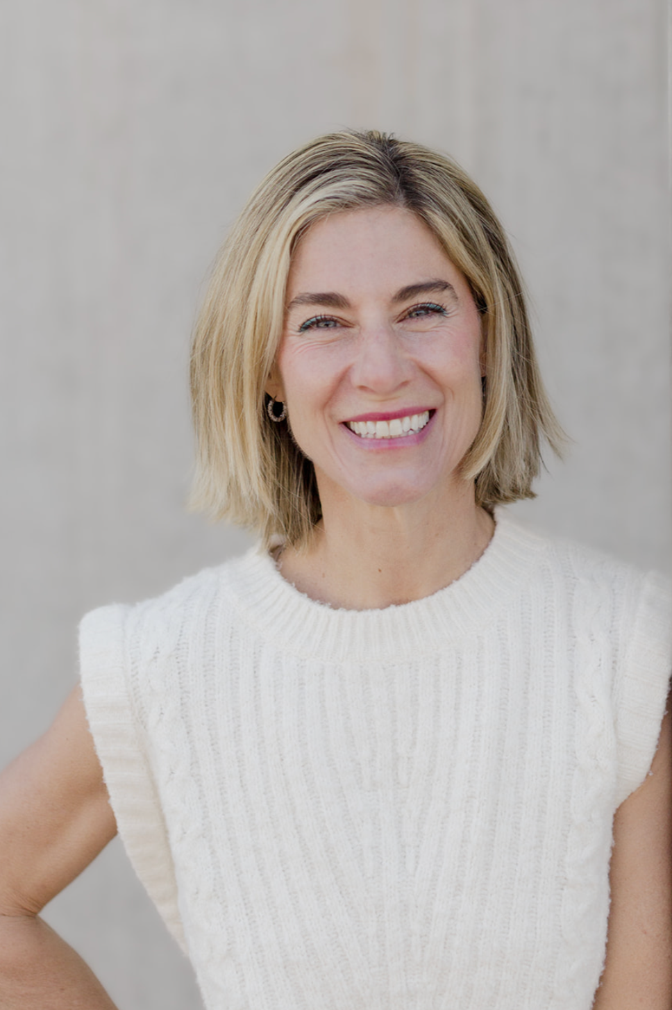 Dr. Kim smiles warmly at the camera, wearing a cream ribbed sweater and hoop earrings, against a soft neutral background.