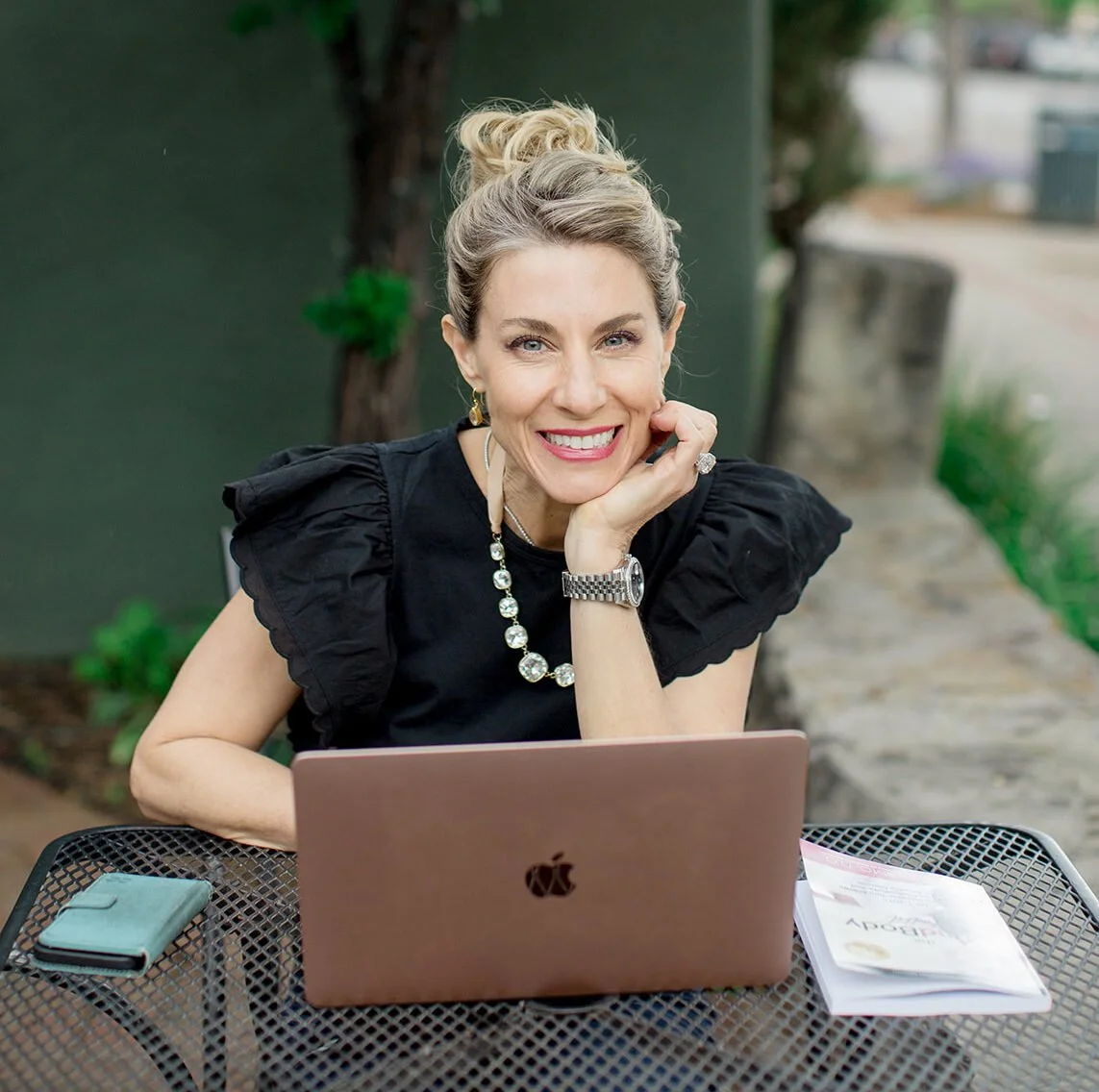 Dr. Kim smiles warmly at the camera, chin resting on her hand, seated at an outdoor table with a rose gold laptop open in front of her.