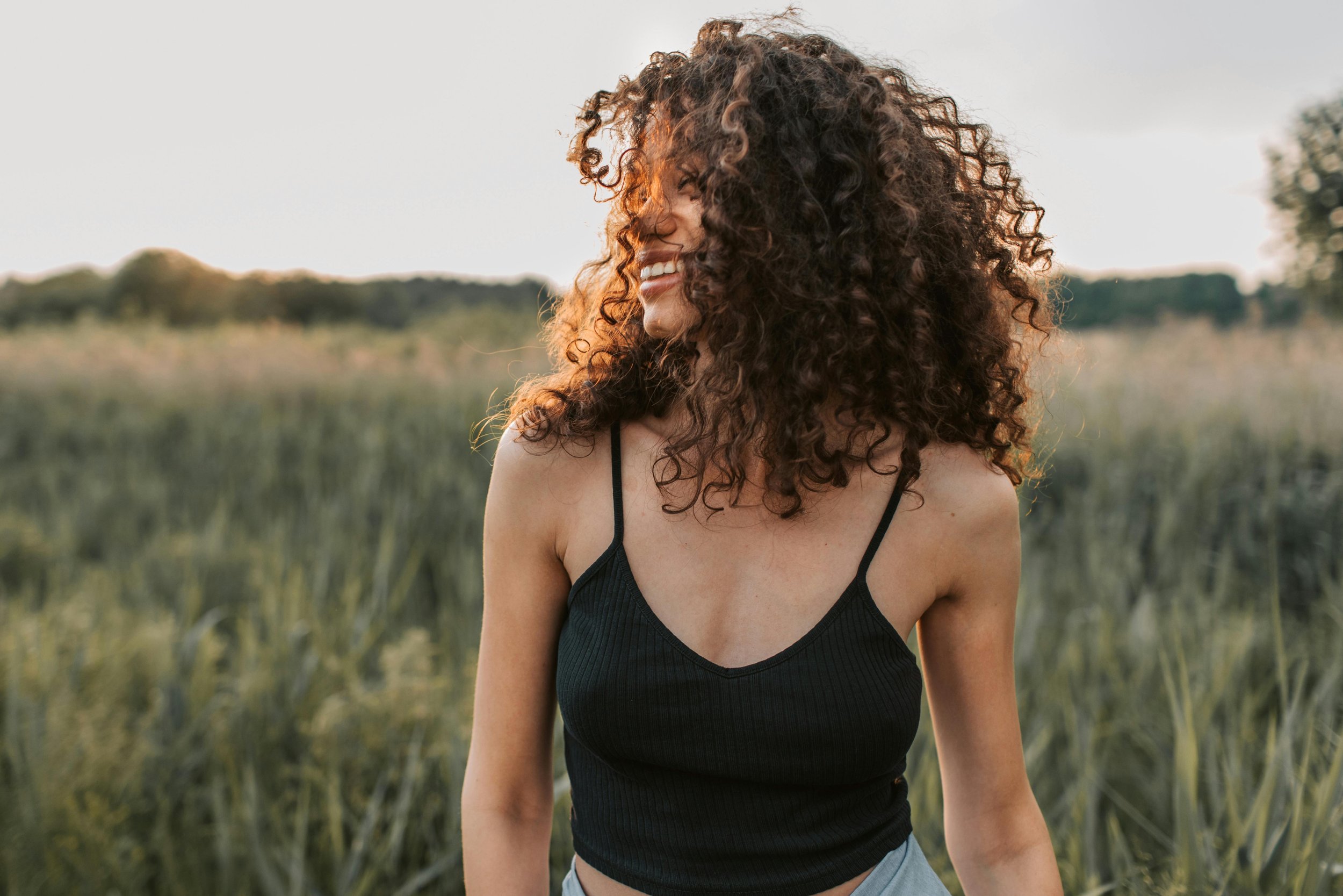 A woman laughs outdoors as wind tosses her curly hair across her face, standing in a sun-warmed field at golden hour.