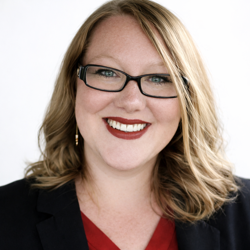 A woman with blonde hair, wearing glasses, red lipstick, earrings, a black blazer, and a red top, smiling at the camera.