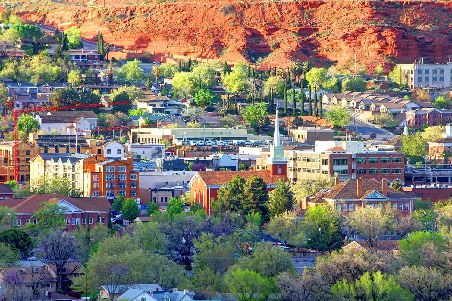 Panoramic view of a small town with a church steeple, various residential and commercial buildings, and a hillside with in the background. Trees and greenery are throughout the town.