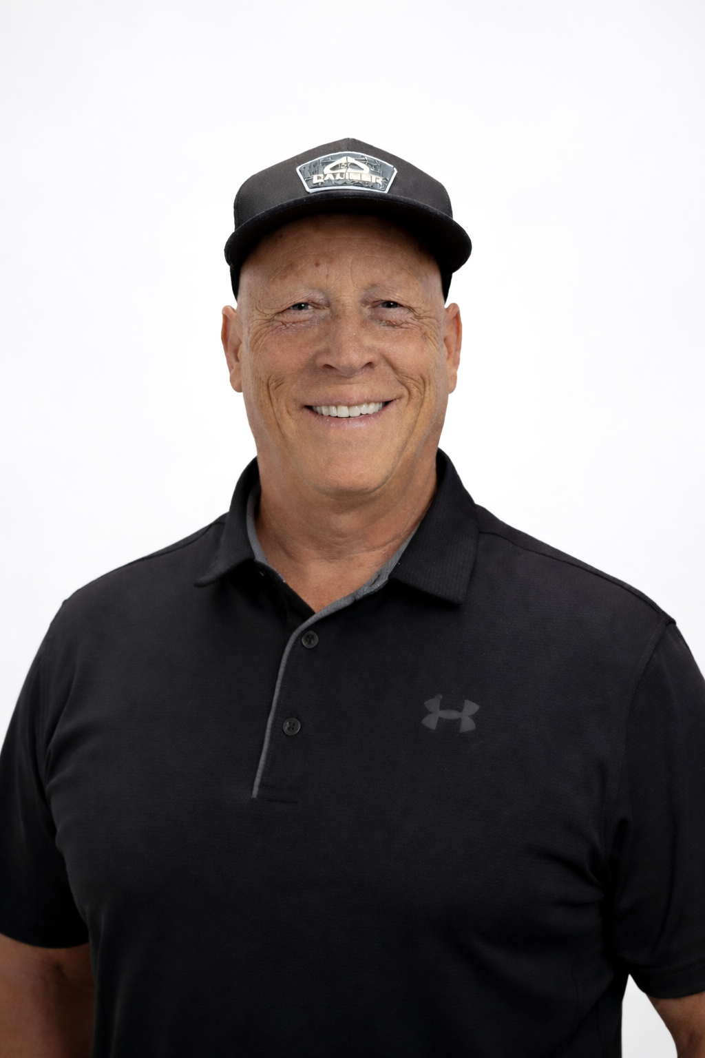A smiling middle-aged man wearing a black Under Armour polo shirt and a black cap with a logo on it.