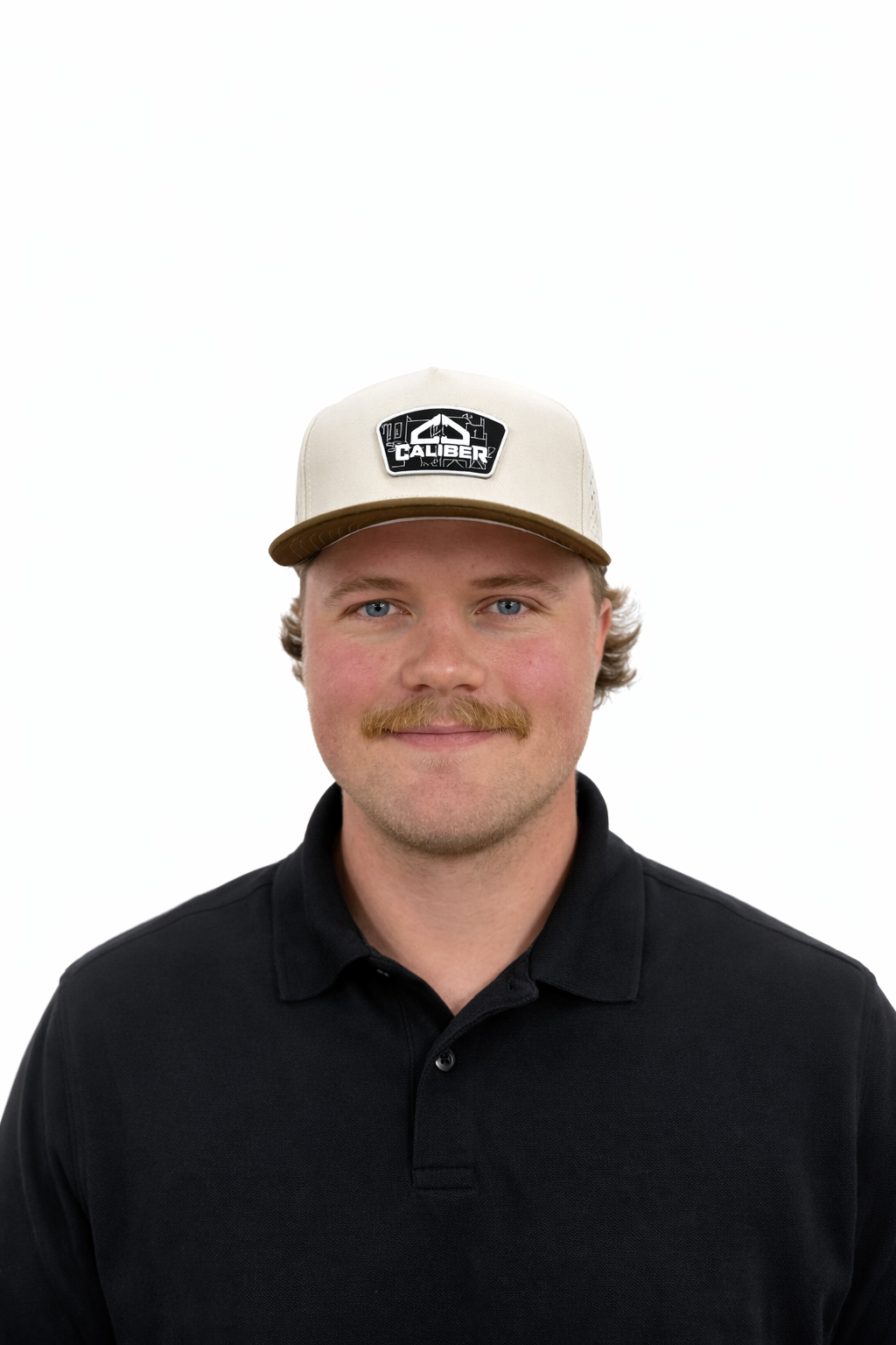 A young man with a mustache and light skin wearing a black polo shirt and a beige and brown cap with a logo on it, smiling at the camera.