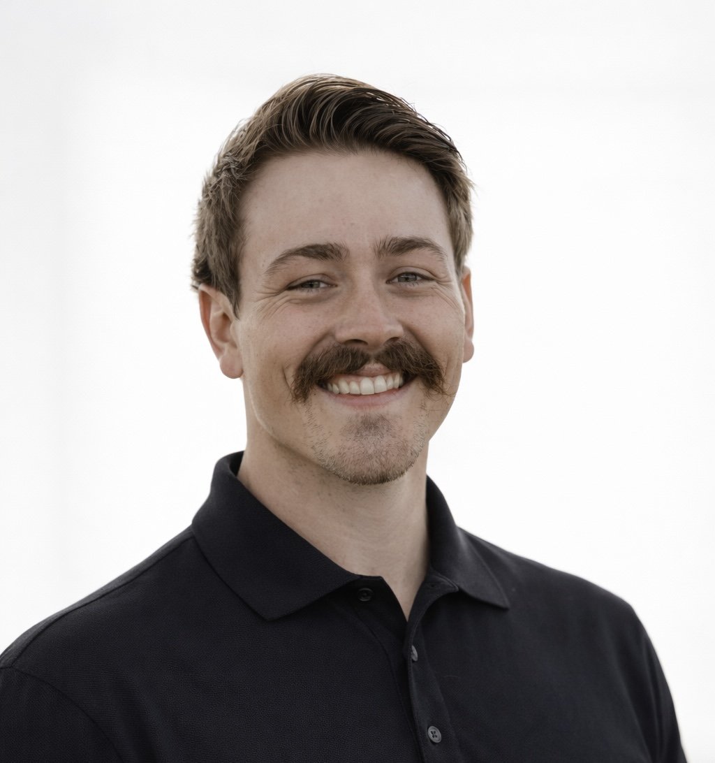 A young man with short brown hair, a mustache, and a beard, smiling and wearing a black collared shirt against a plain white background.
