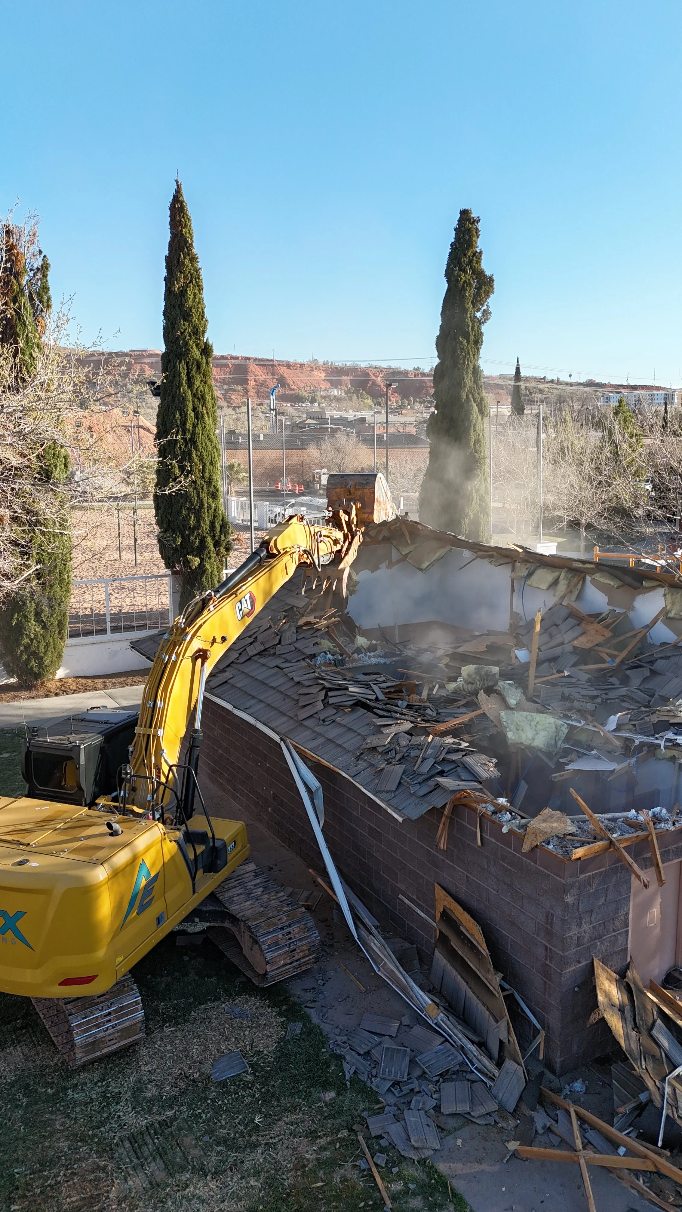 A yellow excavator demolishing a building, with debris and dust in the air, trees in the background, under a clear blue sky.