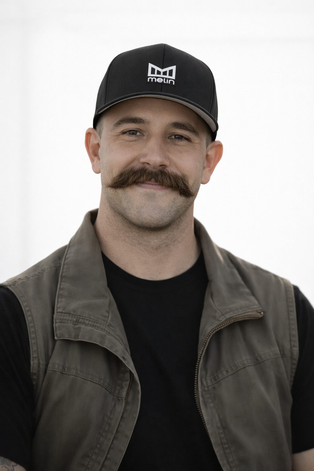 A man with a mustache smiling, wearing a black cap with a logo, a black shirt, and an olive green vest.