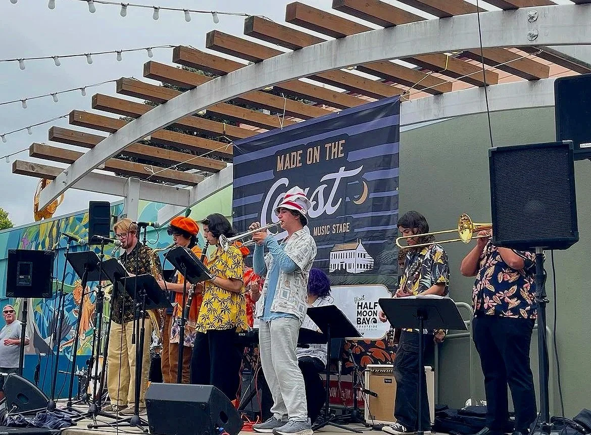 A band of seven musicians performing outdoors on a stage, playing various instruments including a trumpet, saxophone, and keyboard, with a sign that reads 'Made on the Ghost Music Stage' behind them.